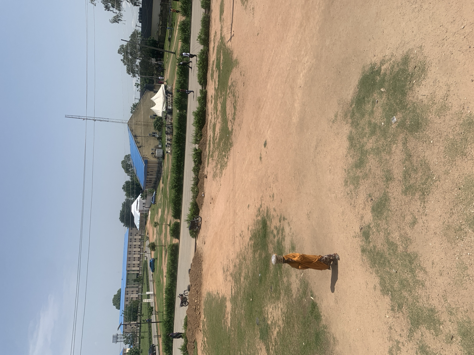 The image depicts an outdoor scene in Jalingo, Nigeria, on a clear, sunny day. The primary subject is a person standing on a dirt and grass area, dressed in a yellow garment and patterned trousers. The person is holding a white object, possibly a cup or container, and is casting a shadow, indicating it is likely midday.
In the background, a paved road runs alongside a neatly trimmed green hedge. Beyond the hedge, there are several buildings with blue roofs, suggesting an institutional or residential complex. A large white tent is visible, potentially for an event. Further back, a tall telecommunications tower pierces the sky, and power lines crisscross the frame. A few cars and motorcycles are parked or moving along the road. The overall setting appears to be a semi-urban environment with a mix of built structures and open, somewhat unkempt land. There is no visible text in the image.