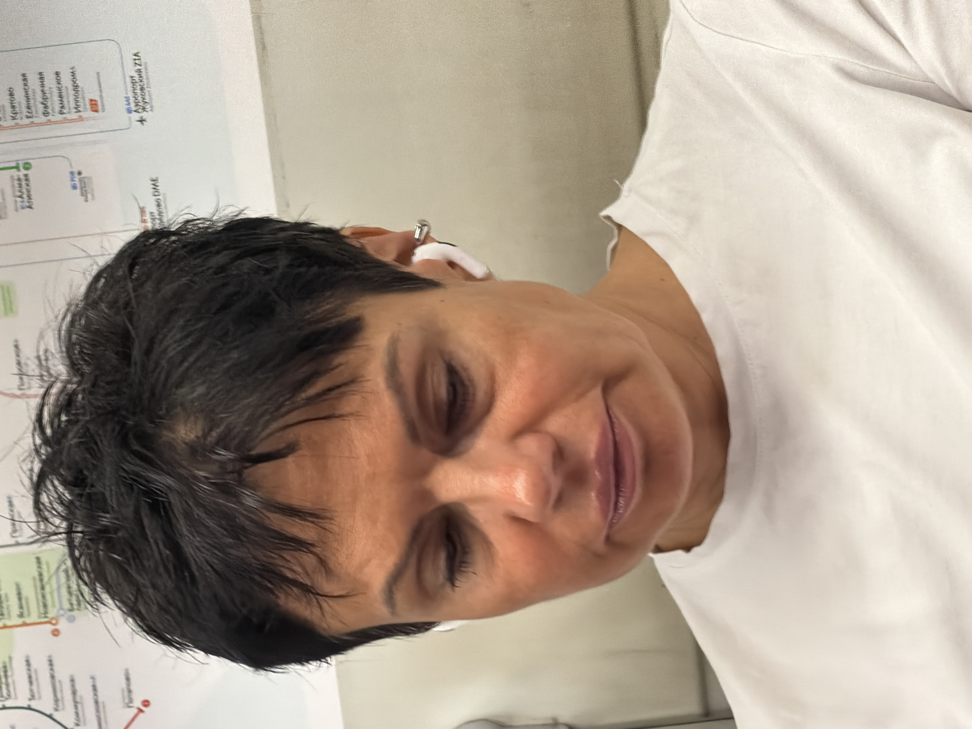A middle-aged woman with short, dark hair and fair skin is shown from the chest up. She is wearing a white t-shirt and white earbuds. She is looking down and to the right. The background appears to be a map or diagram with some text and lines. The woman is in focus, while the background is slightly blurred.