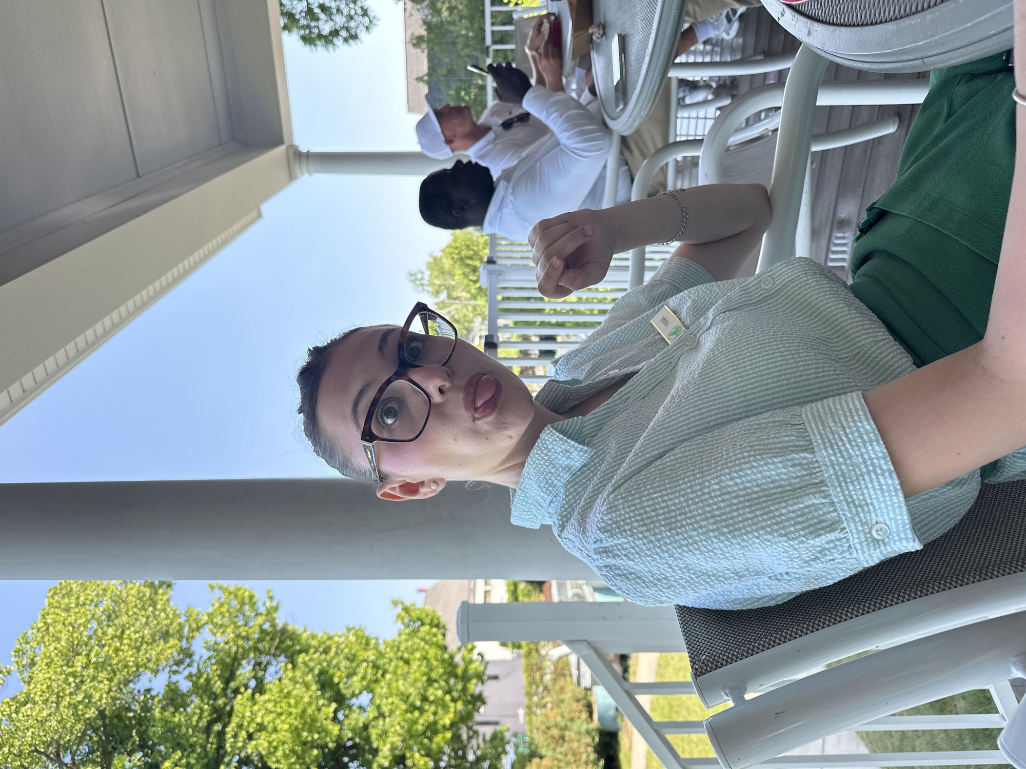 A woman with dark hair pulled back and wearing large framed glasses playfully sticks her tongue out while sitting on a porch in Harbor Springs, Michigan. She is wearing a light blue seersucker button-up shirt and a green skirt. A name tag that reads "YANA" is attached to her shirt. Behind her, two men are seated at tables. The man on the left is wearing a white shirt, and the man on the right is wearing a white cap and shirt, looking at his phone. The porch overlooks a sunny outdoor area with trees and houses in the background. The scene suggests a casual outdoor gathering or meal.