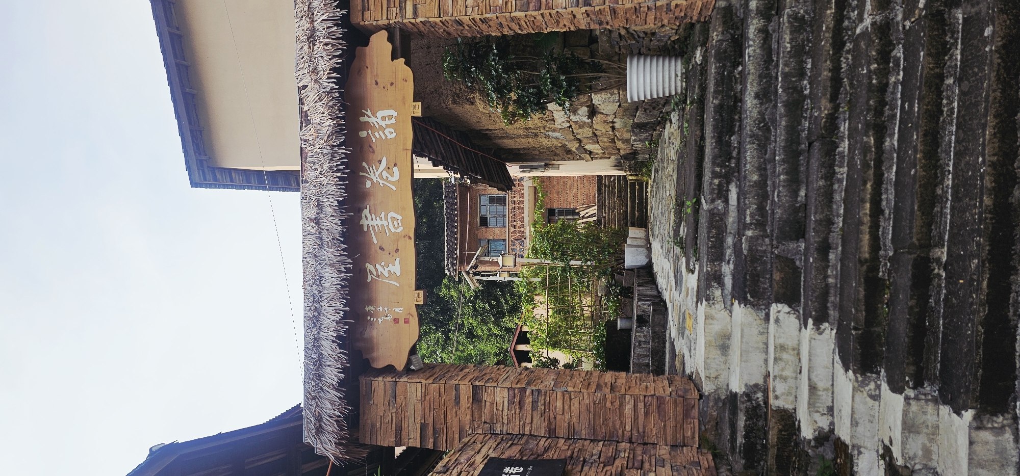 The image shows a pathway leading through an archway into a courtyard area. The archway is constructed with stone pillars and a thatched roof, with a wooden sign inscribed with Chinese characters hanging from it. The characters appear to read "諸卷書屋" (Zhū juàn shū wū), which translates to "Miscellaneous Scrolls Book House".
Beyond the archway, a stone staircase ascends towards a brick building with multiple windows and a decorative balcony. Lush green foliage, including trees and potted plants, surrounds the pathway and courtyard, suggesting a rural or traditional setting. The overall atmosphere is tranquil and hints at a place of learning or cultural significance, possibly a traditional library or a historical site. The location is identified as Shapingba District, China, known for its numerous educational institutions and cultural landmarks.