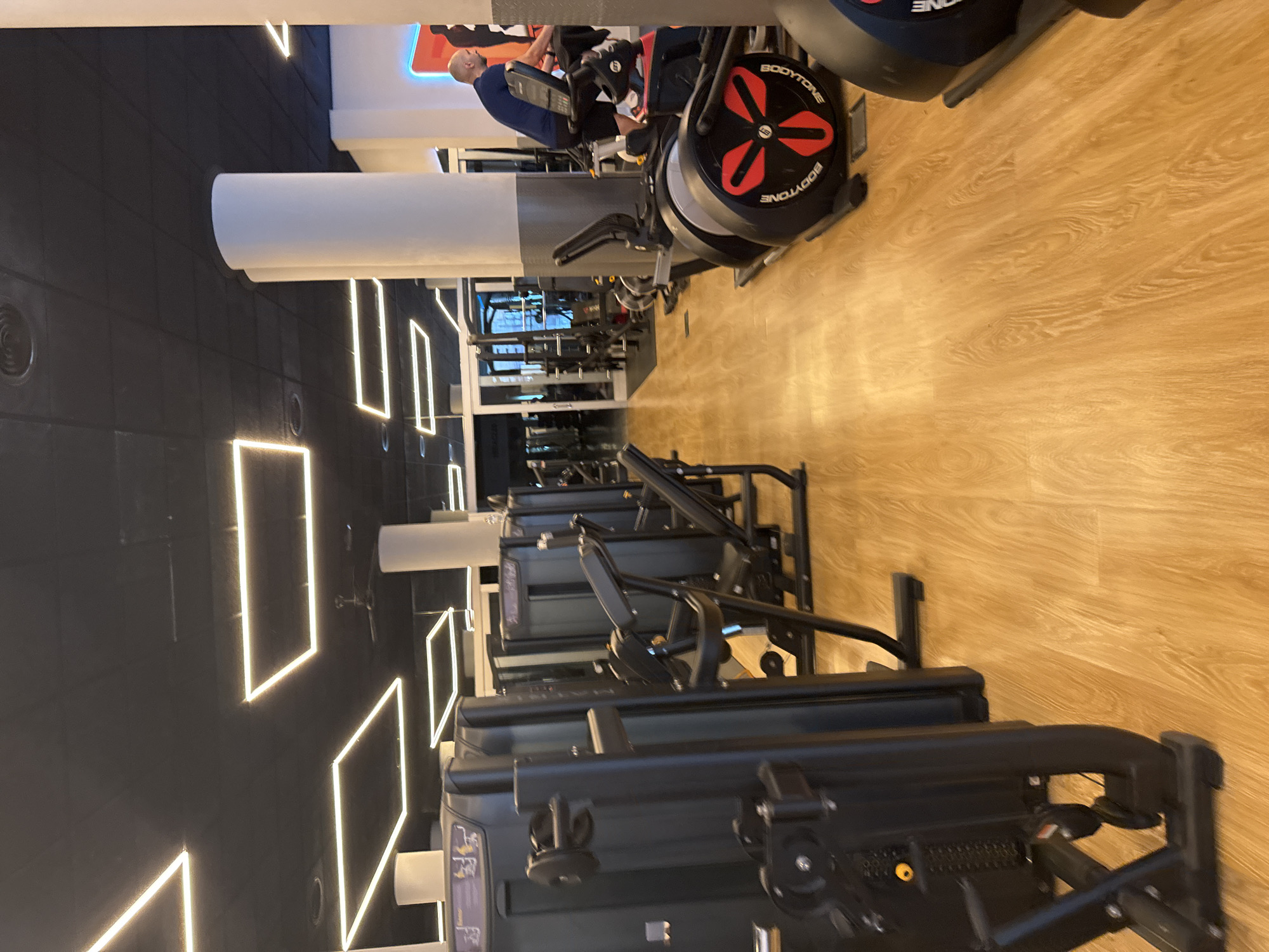 The image depicts the interior of a modern gym located in Murcia, Spain. The gym features a sleek, black ceiling with minimalist, square LED lighting fixtures. The walls appear to be white, punctuated by large, cylindrical white pillars. The floor is a polished light-colored wood.
In the foreground, to the left, is a row of black weight machines, one of which is prominently featured. These machines are part of a gym equipment setup, suggesting a functional workout area.
Further into the gym, in the middle ground, are more weight machines and what appear to be cardio machines, including an elliptical trainer. A man, bald and wearing a dark blue t-shirt and black shorts, is visible on the right side of the image, actively using a stationary bike. He is positioned near the center-right, facing away from the camera and to the left.
In the background, through glass doors, glimpses of other gym areas and possibly outdoor light are visible. The overall impression is a clean, well-lit, and well-equipped fitness facility.