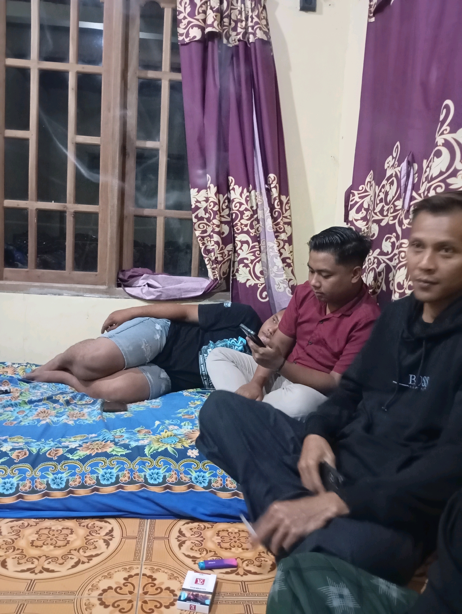 The image depicts a dimly lit interior scene, possibly a living room, in Ketepung, Indonesia. Three men are present. One man in a black t-shirt and denim shorts is lying down on a blue patterned mattress, facing away from the camera. Another man, wearing a maroon shirt, is seated and looking at a mobile phone. The third man, in a black hoodie, is in the foreground, partially obscuring the other two. A wooden window with a lattice is visible to the left, with purple patterned curtains hanging on either side. On the floor, a pack of cigarettes and a vaping device are visible. The overall atmosphere appears casual and relaxed.