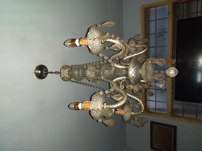 An ornate, multi-armed chandelier crafted from clear glass and metal is suspended from a light-colored ceiling. The chandelier features multiple candle-style light bulbs with golden bases, and numerous decorative glass elements, including teardrop and spherical pendants, are attached to its arms and central column. The ceiling shows minor discoloration around the mounting point of the chandelier. In the background, a light blue-grey wall is visible, with a framed picture hanging on the left side, the details of which are indistinct. A multi-paned wooden-framed window is situated in the lower right and center of the background, below which a dark, rectangular object resembling a television screen is present. The room's specific location within Río Grande, Mexico, cannot be determined from the visual information provided. No individuals are visible, and no actions or interactions are depicted.