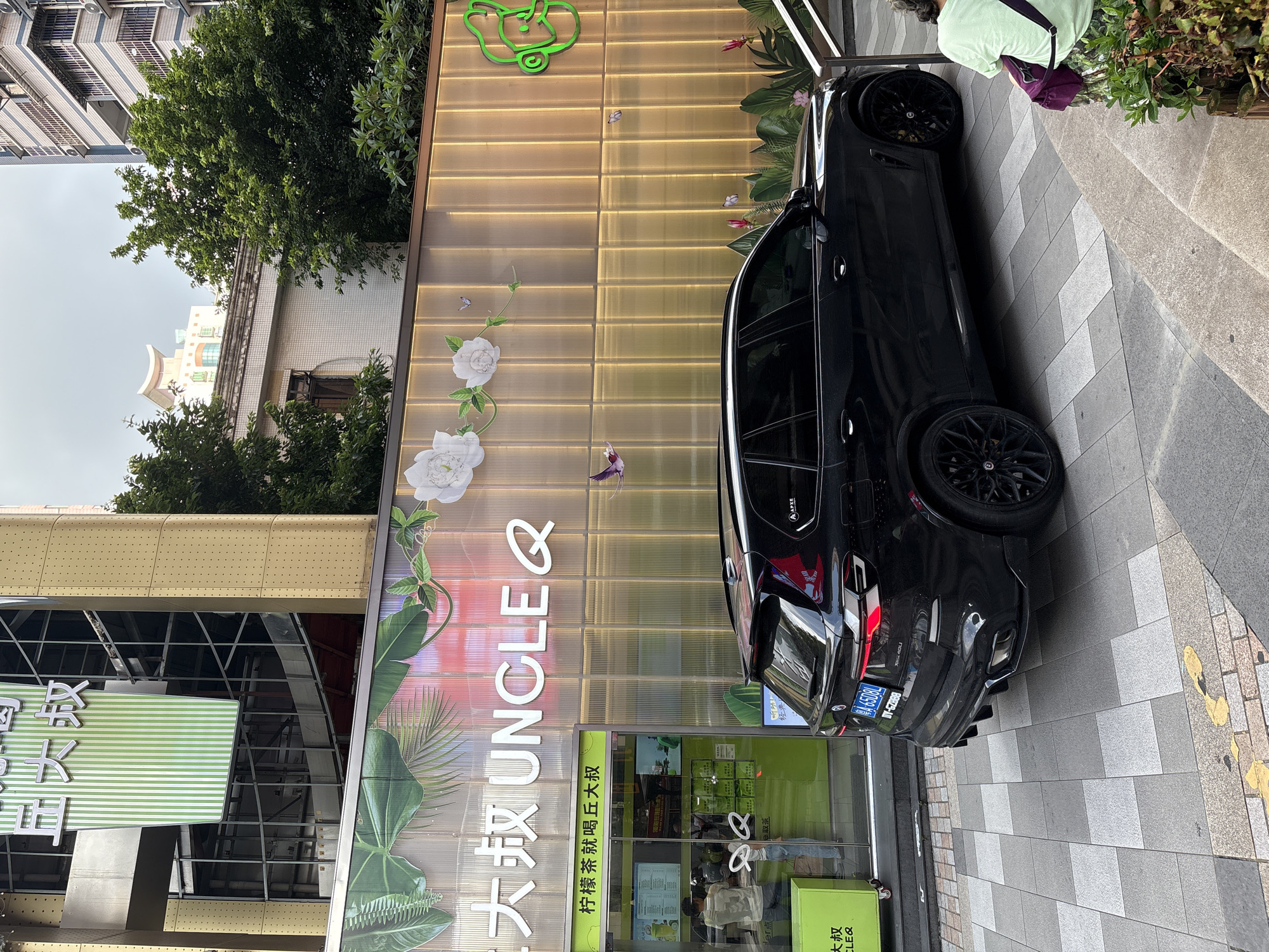 In Haizhu District, China, a black BMW X4 is parked on a tiled sidewalk in front of a store with a bright, golden-hued facade. The store, identified by a sign reading "UNCLE Q" in English and Chinese characters, features large glass windows displaying menus and merchandise. The facade is decorated with images of white flowers and green foliage, and a neon green logo is visible on the right side. To the left of the store, a green cart is partially visible. The street scene is bustling with activity, with a person in a light green shirt sitting on a step to the right of the image, facing away from the camera and holding a purple bag. A building with multiple floors and balconies is visible in the background, along with lush green trees. The sky is overcast.