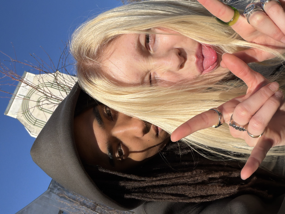 A close-up photograph features two individuals outdoors under a clear blue sky. The background reveals a light-colored building with a distinctive architectural motif of recessed arches and vertical elements, identified as the Palais de Tokyo, located at 13 Avenue du Président Wilson, Paris, France. Bare tree branches are visible against the sky.
On the left, partially obscured by the foreground individual, is a person with dark skin and dark dreadlocks, wearing a dark hooded top. Their face is partially shadowed, and they are looking towards the camera. In the foreground, positioned to the right, is a second individual with fair skin and long, straight blonde hair. This person displays a playful expression, with their tongue extended and their left eye winking. Both of their hands are raised, forming a "rock and roll" or "devil horns" gesture. Several rings are worn on their fingers, including silver bands, a ring with multiple dark stones, a plain gold band, and a translucent green ring. Strong sunlight is evident, casting distinct shadows.