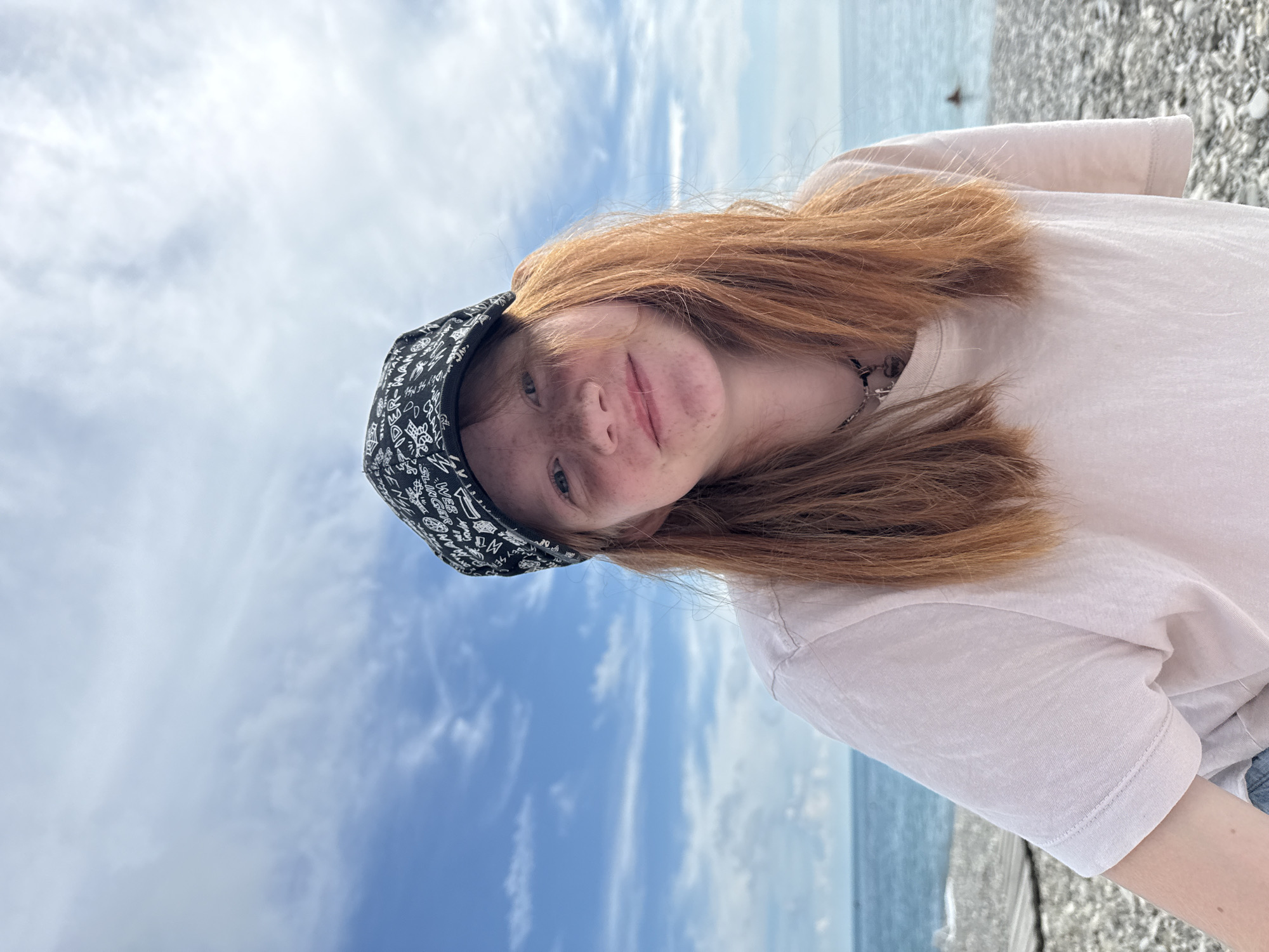 A young woman with red hair and freckles is pictured from the chest up, facing the camera. She is wearing a black baseball cap with a white graphic pattern and a light-colored t-shirt. Her hair is long and falls around her shoulders. She has a necklace with a pendant. The background is a bright blue sky with scattered white clouds. Below the sky is a body of water that appears to be the sea, with a slightly rocky beach in the foreground. The location is identified as Nebug, Russia.