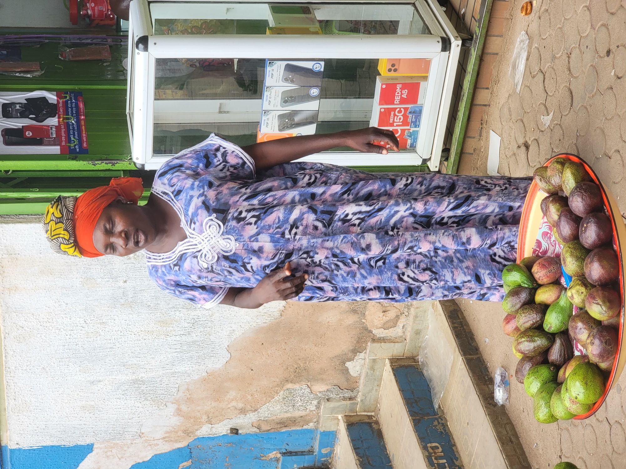 The image, originally rotated 90 degrees counter-clockwise, captures an outdoor market scene in what appears to be Jos, Nigeria. In the foreground, a dark-skinned woman stands, likely a vendor. She is adorned with a colorful orange and yellow headwrap and a patterned blue and white traditional dress. Her right hand is casually placed near a white-framed glass display case.
To the woman's right, a large orange tray is filled with numerous avocados of varying ripeness, some green and some with a dark purplish hue. This suggests she is a fruit seller. Behind her, a green wall serves as a backdrop, featuring various advertisements and phone boxes. The glass display case prominently showcases mobile phones, with visible branding for "TECNO POP 10," "TECNO POP 10 Pro," "REDMI 15C," and "REDMI A5."
A promotional sign on the green wall announces: "We are giving out 3 Phones Every Week." Further text encourages customers to "verify your phone's authenticity to enjoy this benefit," with an offer "valid till 08 November 2023." The ground is paved with light-colored bricks, showing some wear, and a blue-painted step or curb is visible. A discarded plastic bag and paper are on the pavement. The scene is brightly lit, indicating a daytime setting. The woman appears engaged in her activity, contributing to the dynamic atmosphere of a local market.