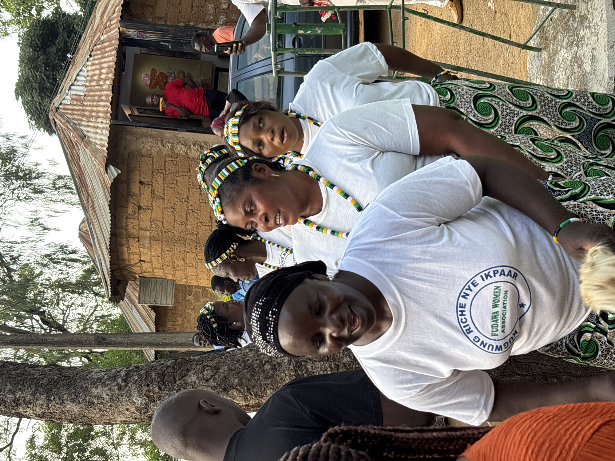 The image depicts an outdoor scene in Jos, Nigeria, with a group of women in the foreground wearing white t-shirts and traditional patterned skirts. They are adorned with beaded necklaces and headbands in green, yellow, and black. One woman in the immediate foreground is wearing a black headscarf decorated with silver sequins. Behind the women, a rustic building with a corrugated metal roof is visible. A man in a red shirt is standing in the doorway of this building. Further in the background, a car and a fence are present. The overall atmosphere appears to be a community gathering or event.