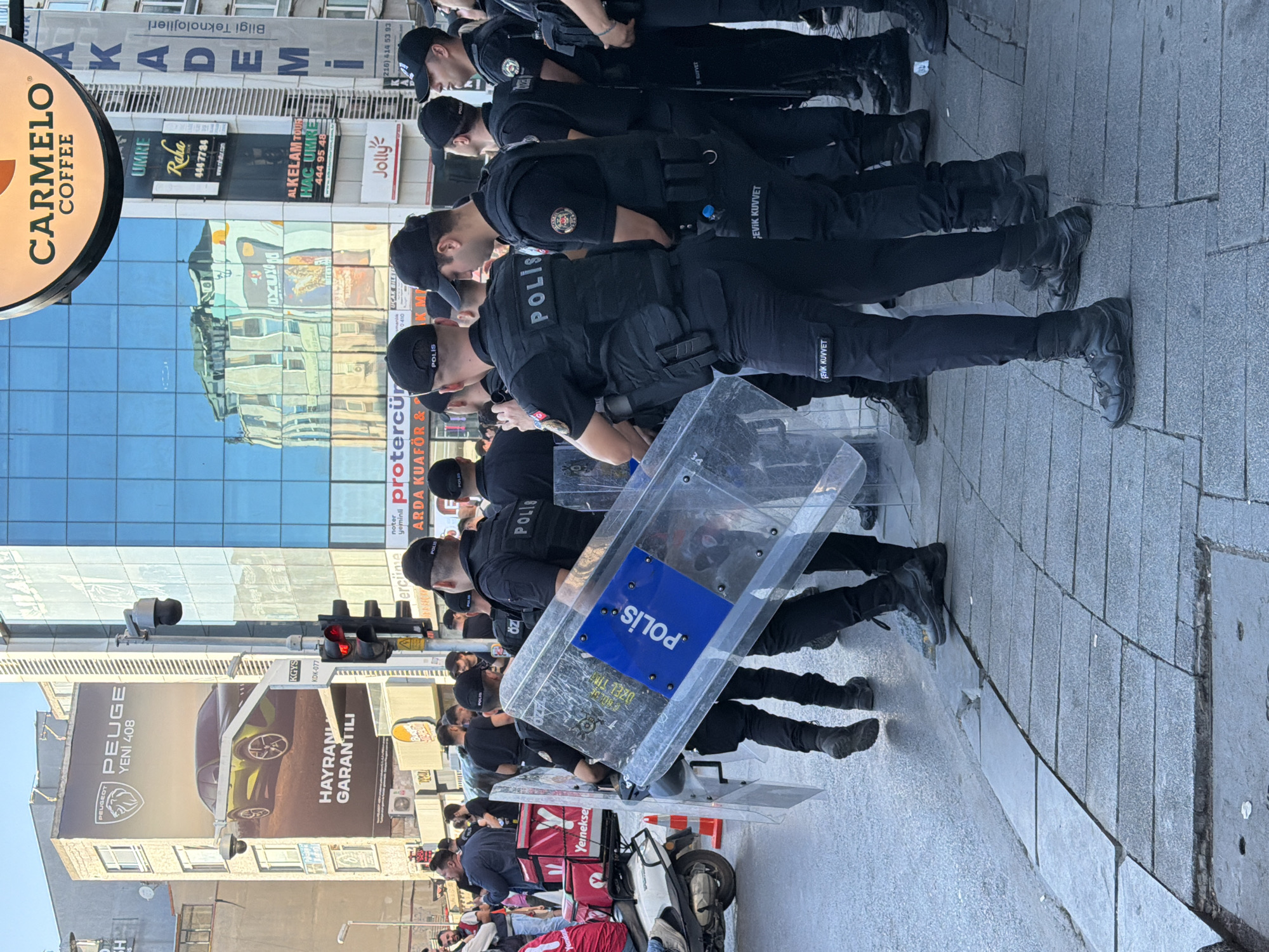 In Kadıköy, Turkey, a group of police officers in black uniforms stand in formation on a city street. They are equipped with transparent riot shields. The officers are positioned in front of a building with a blue glass facade, reflecting the surrounding environment. On the building's exterior, advertisements are visible, including one for "CARMELO COFFEE" and another for "PEUGEOT YENI 408". A traffic light is present, indicating an intersection. The ground is paved with stone slabs. Some officers are holding their shields, while others have them lowered. The overall scene suggests a law enforcement presence in a public urban area.