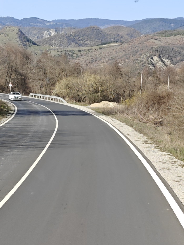 A two-lane asphalt road, marked with a solid white line on the right edge and a dashed white center line, curves through a rural, mountainous landscape in the vicinity of Gospodintsi, Bulgaria. The road initially bends sharply to the left, where a white four-door sedan is observed driving towards the viewer. A metal guardrail borders the left side of the road on this curve, accompanied by a triangular warning sign on a pole. The road surface exhibits dark tire marks and appears recently paved or well-maintained.
To the immediate right of the road is a narrow gravel shoulder, transitioning to dry grass and sparse, leafless deciduous vegetation, consistent with a late autumn or early spring season. Further along the right embankment, a pile of light-colored earth or sand is visible, along with two thin white poles. The background consists of multiple layers of undulating hills and mountains, covered predominantly by leafless trees and some patches of lighter vegetation. The sky overhead is clear and light blue. Sunlight casts long shadows across the road from the left, indicating a low sun angle. No individuals are present in the visible scene.