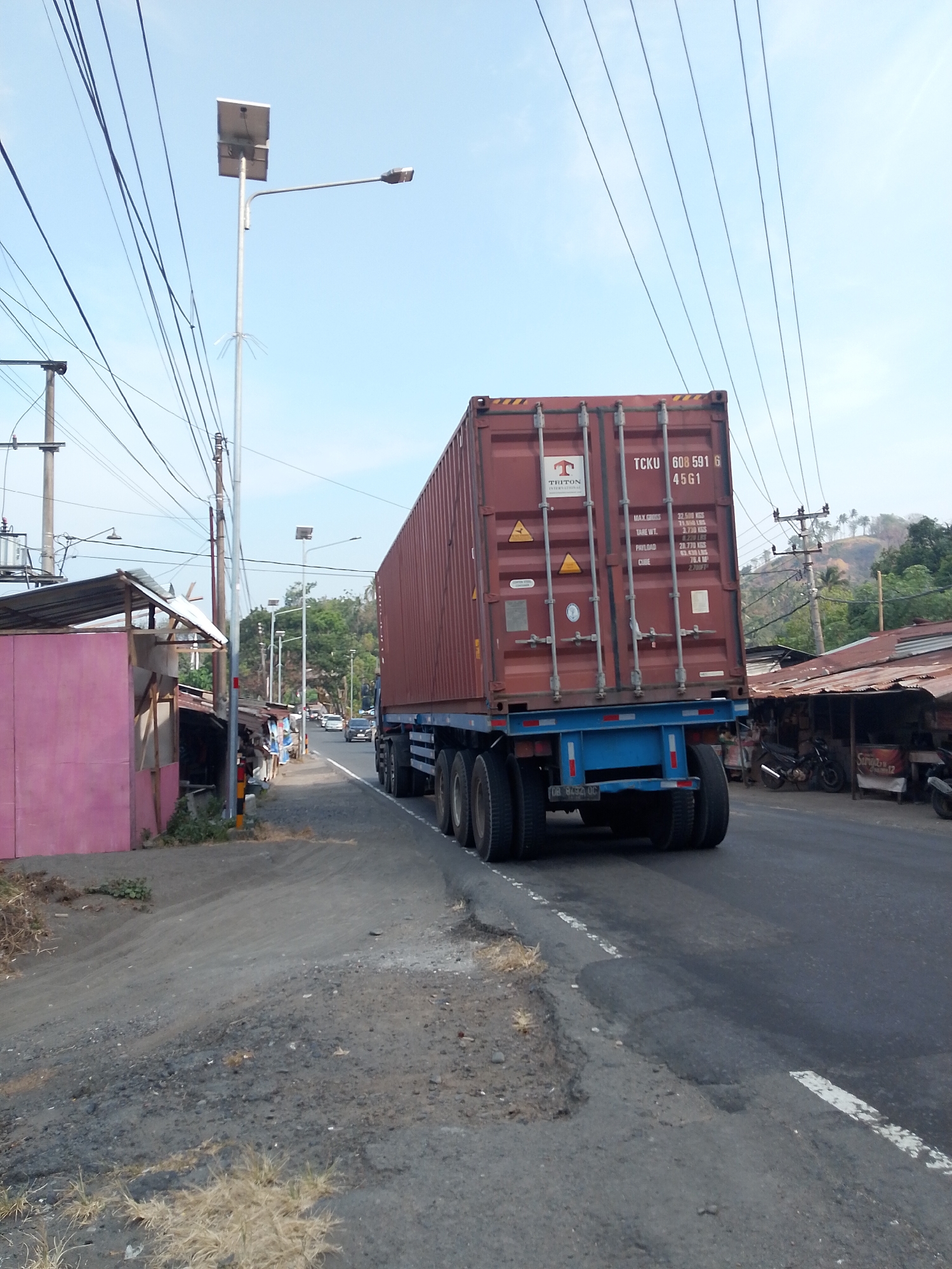 This image depicts a scene on a road in the city of Teep, Indonesia. The main subject is a large, red shipping container being transported on a trailer by a truck. The container is marked with "TTCKU 608 591 6 45G1" and "TRITON". The truck has dual rear wheels and is driving on an asphalt road. To the left of the road, there is a pink building with a corrugated metal roof, and next to it, a cluster of makeshift structures with corrugated metal roofs. Further down the road, several vehicles, including cars and motorcycles, are visible, and there are more small structures and trees. In the background, a hillside with trees and a power line with a street light is present. The sky is clear and blue. The overall environment suggests a semi-urban or rural roadside area.