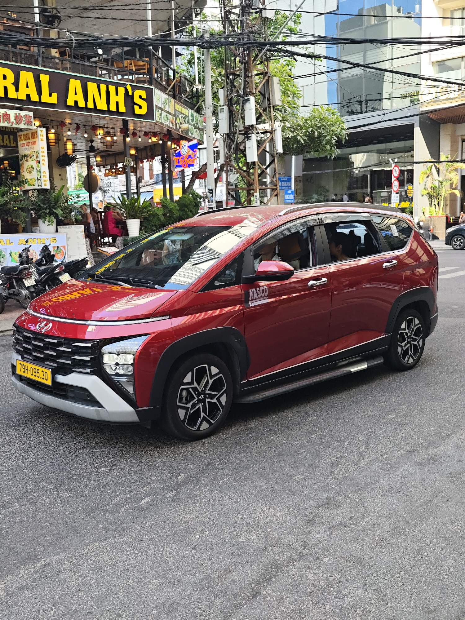 This is a street-level photograph taken in Nha Trang Ward, Vietnam, showing a red Hyundai Stargazer MPV driving on an asphalt road. The car has a license plate that reads "79H-095.30" and features a black "NASCO" logo on its side. The vehicle is captured in motion, with its wheels slightly blurred.
In the background, a building with a prominent yellow sign displaying "RAL ANH'S" is visible. The building appears to be a restaurant or shop, with outdoor seating and potted plants. Several motorbikes are parked in front of the establishment. Overhead, a complex network of electrical wires and utility poles crisscrosses the sky, indicative of an urban environment. The overall scene depicts a typical streetscape in a Vietnamese city, with a focus on the car and its immediate surroundings.