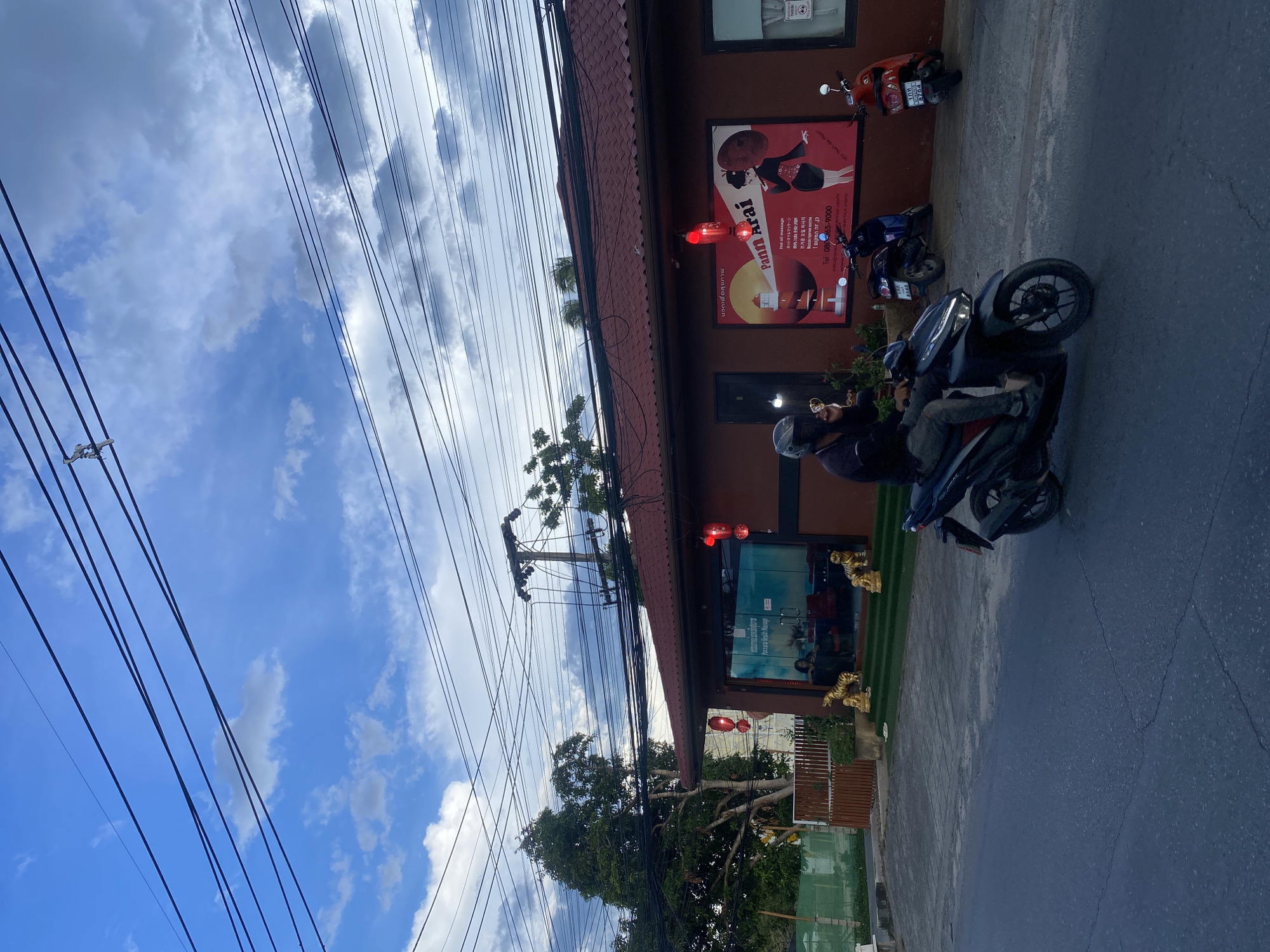 The image shows a street scene in บ้านบางรักษ์, Thailand. A man on a black and gray motorcycle is parked outside a business. The business has a red awning and a sign with the words "Pattarai Health Massage" written in multiple languages. There are two small red lanterns hanging outside. Two other motorcycles are parked nearby. The sky is blue with white clouds. A network of power lines is visible overhead. There are some trees and greenery near the business.