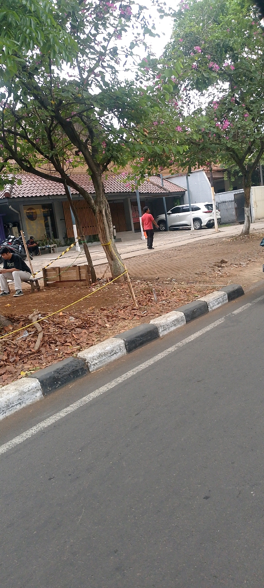 The image depicts an outdoor scene in Bandung City, Indonesia. In the foreground, a paved road runs diagonally from the bottom left to the right, bordered by a curb with alternating black and white painted sections. Beyond the curb, the ground is covered with dry leaves and dirt. A wooden bench sits to the left, with a young man in a black t-shirt and light-colored pants sitting on it and looking at his phone. Behind him, to the left, is a structure with a tiled roof.
Further back, a person in a red t-shirt and black pants is walking away from the camera towards a white SUV parked under a covered area. Trees with green leaves and some pink flowers are scattered throughout the scene, casting shadows. The background reveals buildings with tiled roofs and a fence. The overall impression is that of a street scene in a residential or commercial area, possibly with some ongoing development or renovation, given the presence of yellow caution tape.
