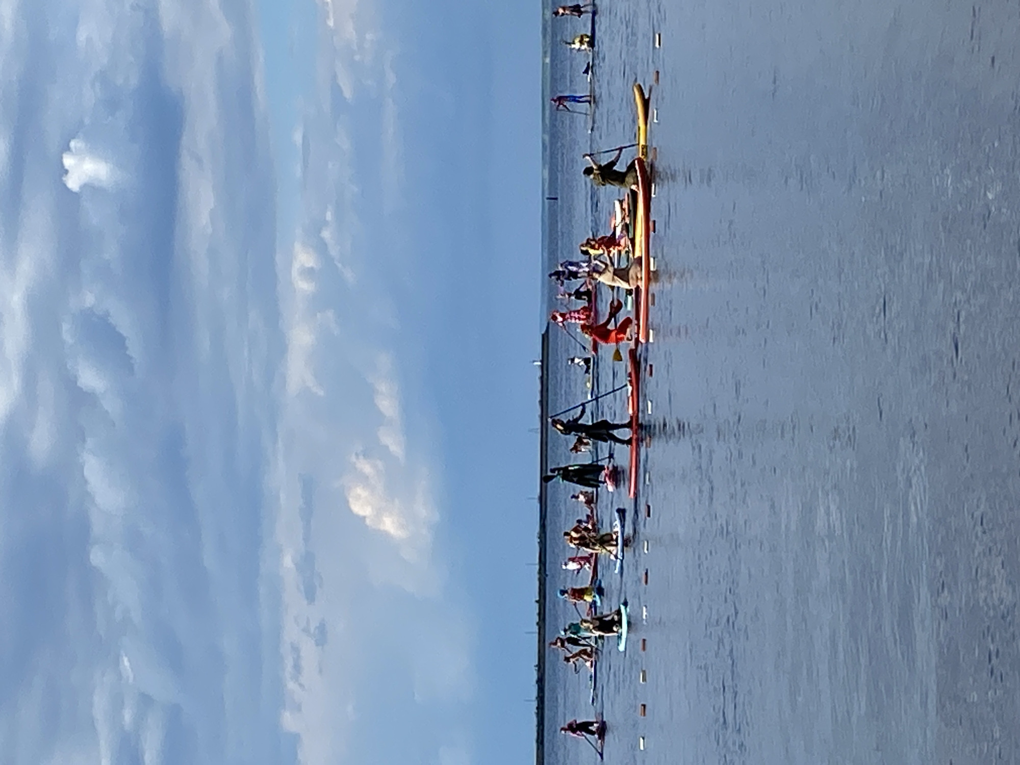 The image depicts a group of people participating in a paddleboard race on a body of water. Several individuals are on paddleboards, some standing and actively paddling, while others are seated. The race appears to be underway, with participants spread out across the water. The setting is outdoors, with a clear blue sky featuring scattered white clouds. The water is calm and reflects the sky. The scene suggests it is daytime, likely late morning or afternoon, given the bright lighting. The image was taken in Chaikovsky, Russia. There are no visible signs or text in the image. The overall mood is one of athletic competition and outdoor recreation.