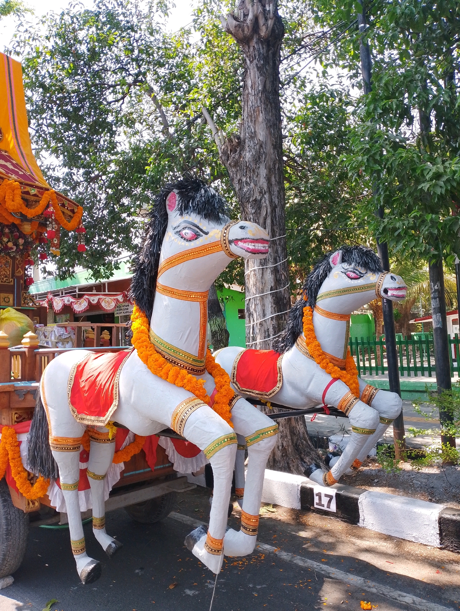 A brightly decorated float with two life-sized horse effigies is parked outdoors, likely on a street in Probolinggo, Indonesia, as suggested by its context. The float, adorned with orange flower garlands, red saddle blankets, and gold trim, is hitched to a vehicle, partially visible on the left. The horses are white with black manes, exaggerated red and pink eye makeup, and open mouths showing red tongues. They are decorated with similar orange garlands and golden bands around their legs. Behind the float, lush green trees form a canopy, with a tall tree trunk prominently visible in the center. The ground is paved with asphalt, marked with white and black curbs, and a white number "17" is affixed to the curb. The scene suggests a festive or ceremonial occasion, possibly a parade or religious procession.