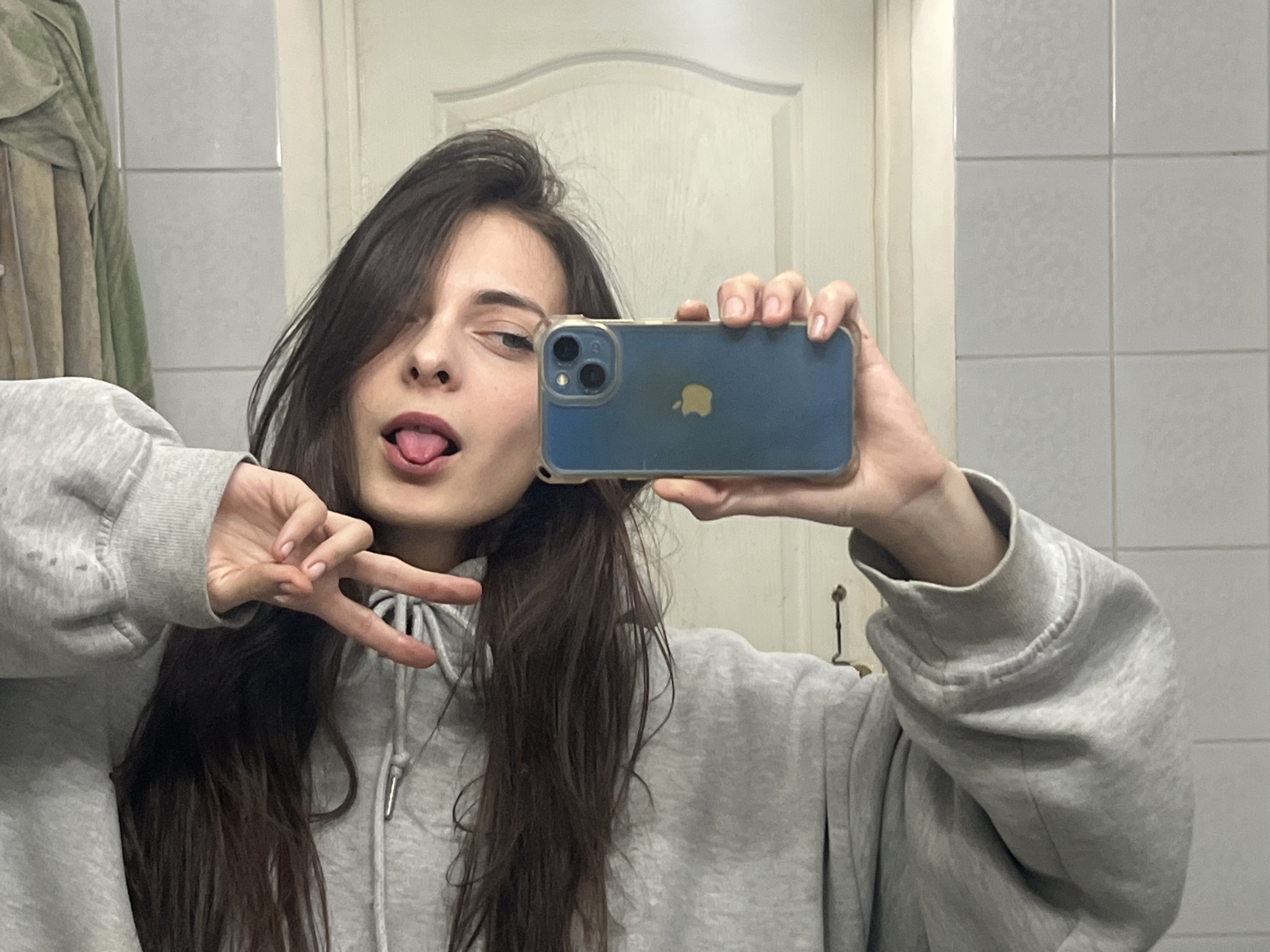 A young woman takes a selfie in a bathroom, presumably in Krasnoyarsk, Russia, based on the metadata. She wears a grey hoodie and has long brown hair. Her tongue is sticking out, and she winks while holding up a peace sign with her left hand. She holds an iPhone with a blue case in her right hand. The background shows white tiled walls and a white door with a raised panel design. A partial view of what appears to be a garment hangs on the left side of the frame.