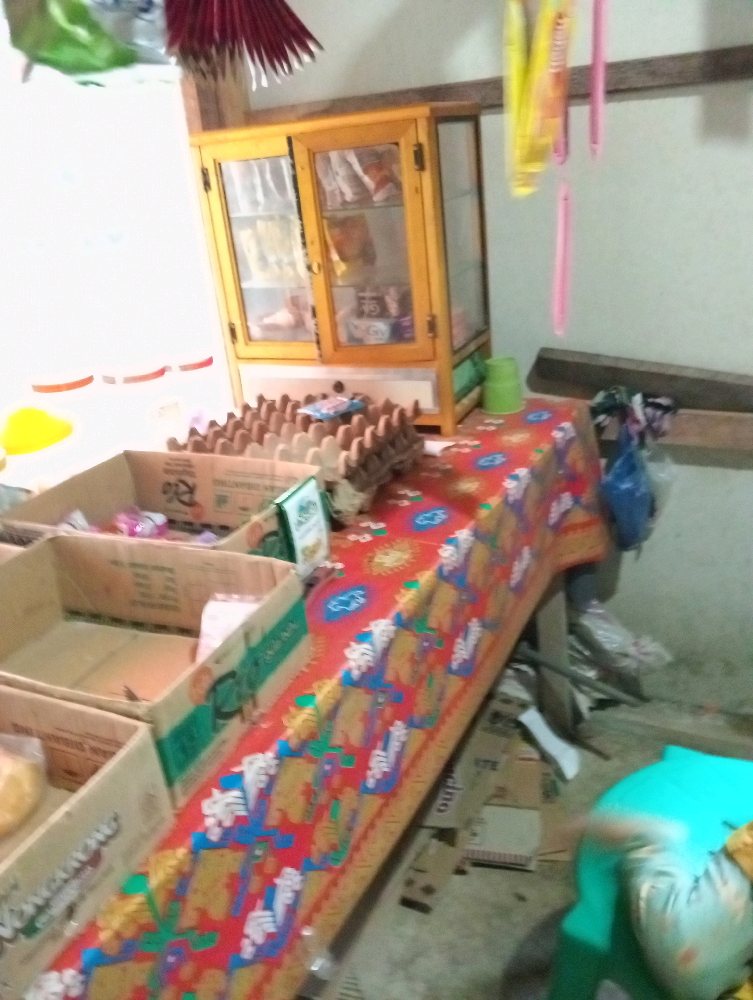 In Campago Barat, Indonesia, a dimly lit room appears to be a small shop or market stall. A wooden cabinet with glass doors stands against a wall, displaying various items. In front of the cabinet, a long table is covered with a red cloth featuring a colorful, patterned design. On the table, there are several cardboard boxes, some containing items, and a tray filled with eggs. To the right of the table, there are more boxes and bags on the floor. A bright turquoise bag is visible in the lower right corner. The overall impression is of a modest, cluttered retail space.