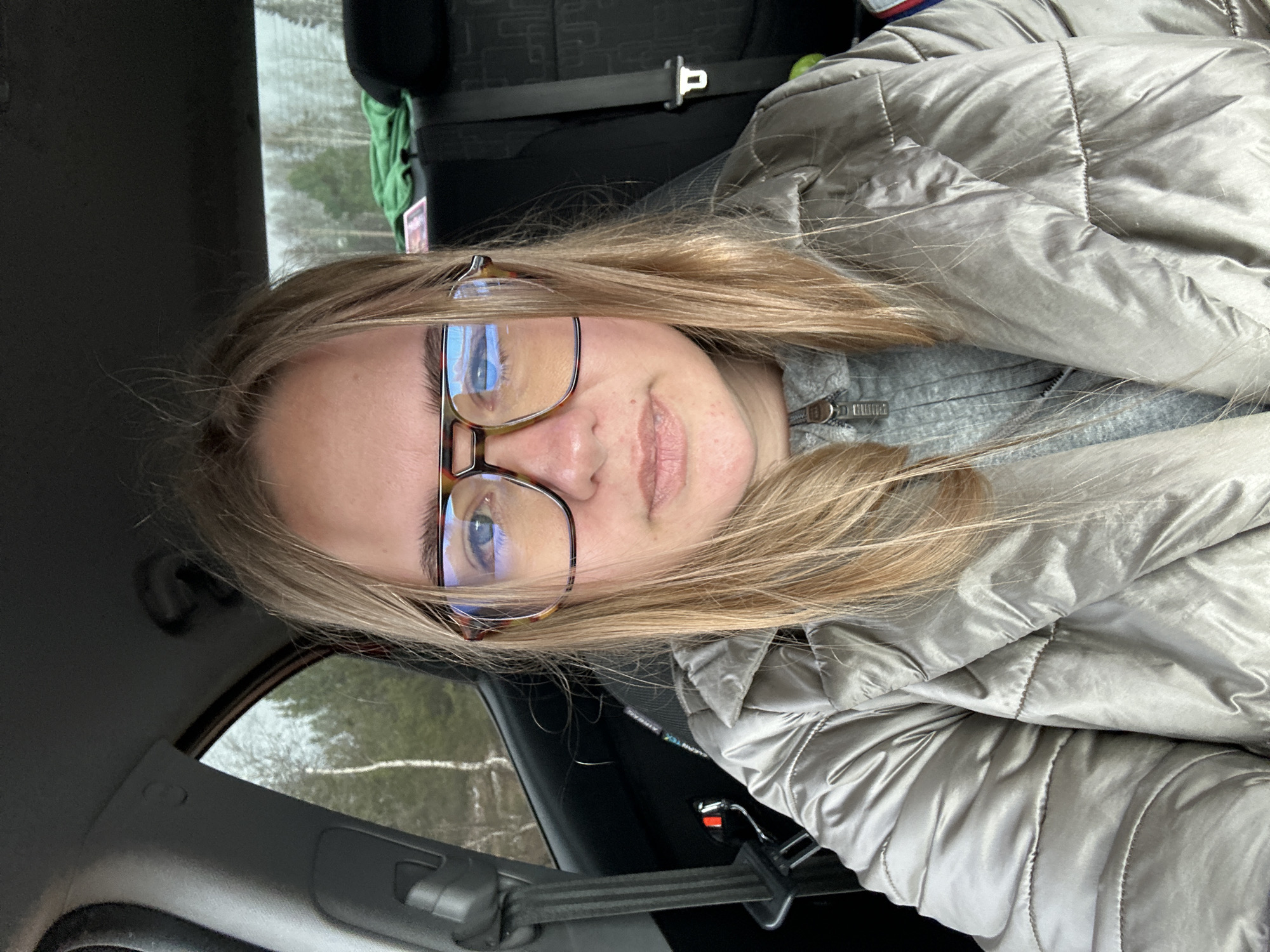 A young, fair-skinned person with blonde hair and glasses is seated in a car, likely in the driver's seat, as indicated by the seatbelt. The person is wearing a grey, quilted jacket over a grey hooded sweatshirt. The interior of the car is visible, with the car window in the background showing a landscape with trees, possibly indicating a road trip. The photo does not contain information to confirm the location to Ural'skoe, Russia.