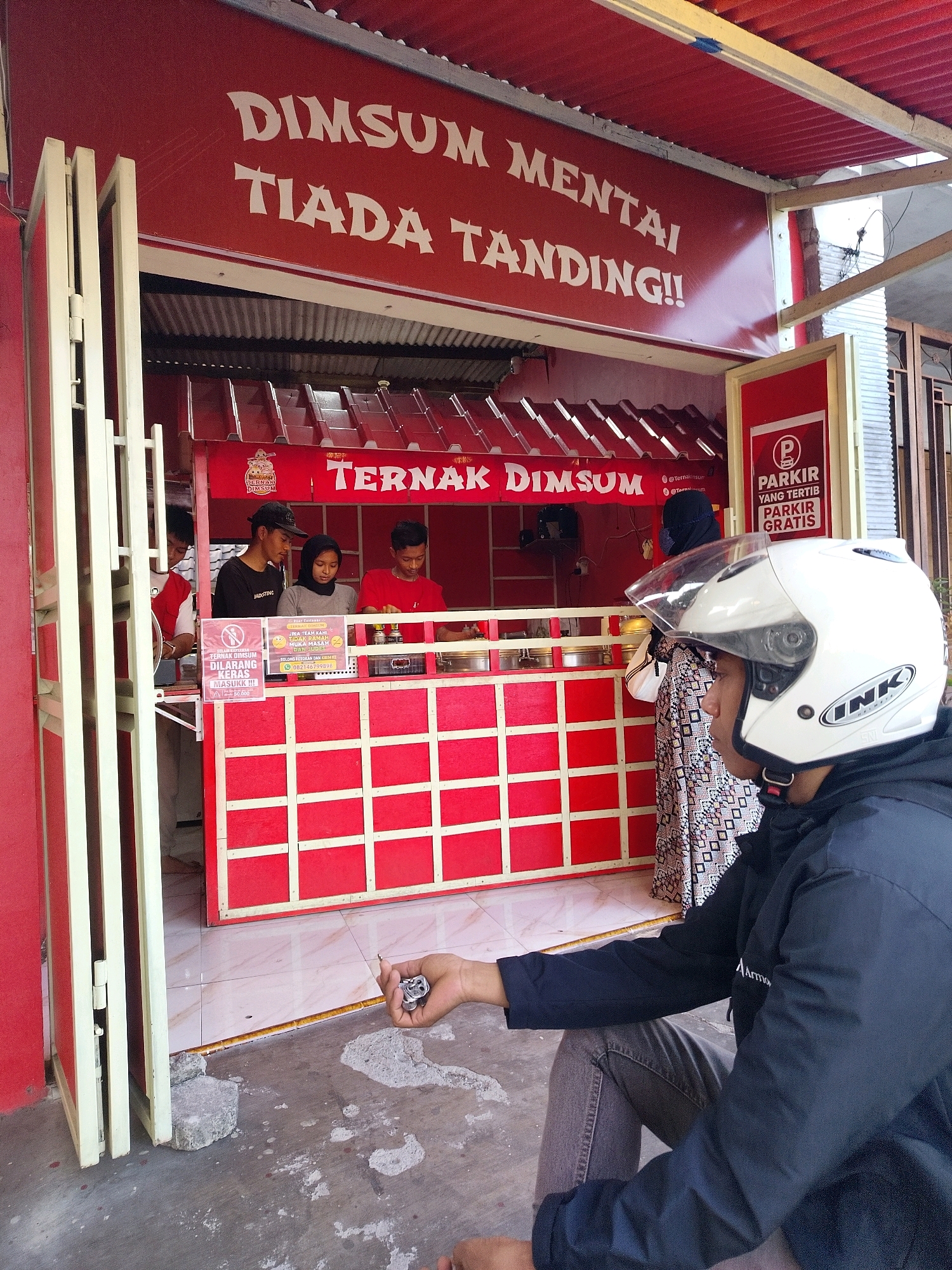 The image depicts a red food stall with white lettering that reads "DIMSUM MENTAI TIADA TANDING!!" in the upper section and "TERNAK DIMSUM" below. A group of people are visible behind the counter. On the left, a person wearing a red and white shirt appears to be working. Next to them is a person wearing a black cap and t-shirt. In the center, a person in a gray hijab and t-shirt is looking downwards. To the right, a person in a red t-shirt is also behind the counter. A sign on the counter indicates "DILARANG KERAS MASUKK!!!" and has a phone number. Another sign on the right side of the stall displays a parking symbol and text that translates to "PARKING FOR FREE". In the foreground, a person wearing a black jacket and helmet is seated, facing away from the stall and looking towards the viewer. The stall is situated in an outdoor environment, possibly a street or market, with a tiled floor and a concrete ground. The overall scene suggests a commercial transaction at a dim sum stall in Mojokerto, Indonesia.