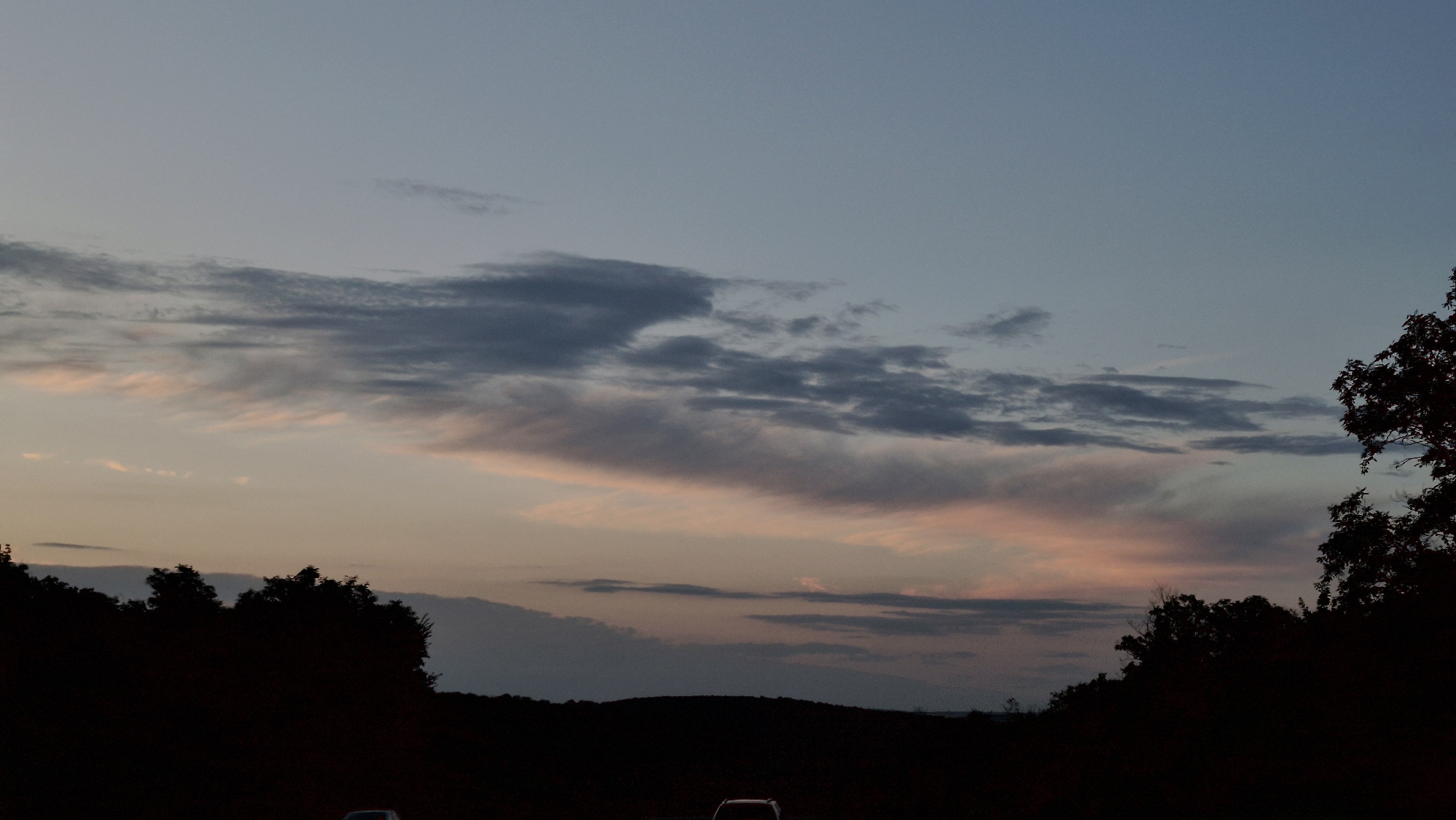 The image captures a twilight scene with a serene sky filled with wispy clouds tinged with pink and orange hues. The horizon is silhouetted by a line of trees and undulating hills, suggesting a rural or suburban landscape. In the foreground, the faint outlines of two vehicles are visible, indicating a road or parking area. The lighting and composition suggest a peaceful, transitional moment between day and night. The location of Razeni, Moldova is not explicitly depicted or inferable from the visual information provided.