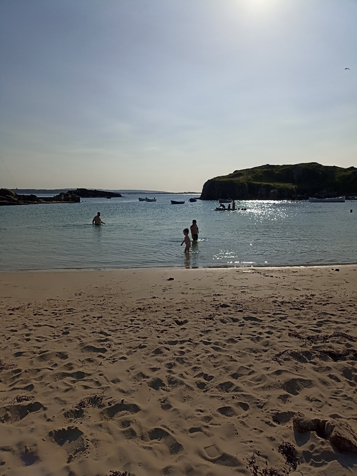 The image captures a serene beach scene at Carnboy, Ireland, under a bright, hazy sky. The foreground is dominated by a sandy beach, showing subtle undulations and scattered seaweed. Gentle waves lap at the shore, leading into clear, shallow water. Several people are visible in the water, including two young children near the shore and an adult further out. In the mid-ground, a rocky island or promontory is silhouetted against the bright sky, with a few boats anchored nearby. The water reflects the sunlight, creating a shimmering effect. The overall atmosphere is one of a peaceful summer day at the coast.