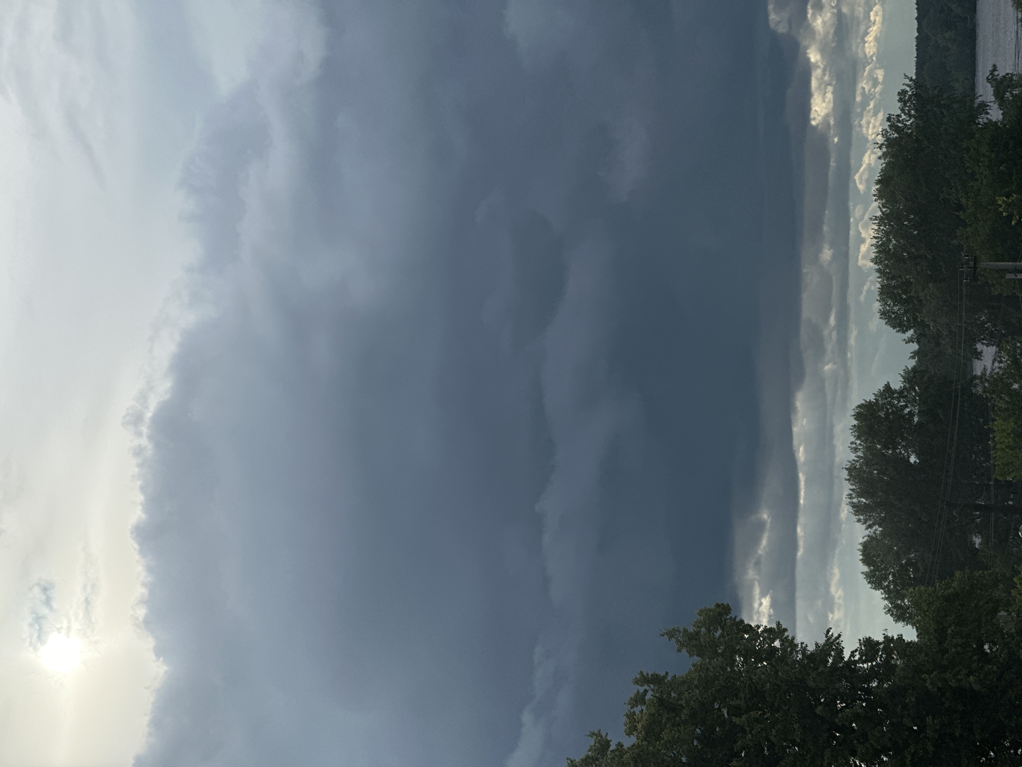 The image presents a dramatic natural scene dominated by a contrasting sky, suggesting a developing or approaching storm. The frame is divided by a distinct vertical line of clouds. On the left side, the sky is bright and partly cloudy, with the sun partially visible through lighter clouds, casting a soft, diffuse glow.
The right side of the image is filled with a vast, dense mass of dark, grey-blue storm clouds, indicating heavy weather. This dark cloud mass extends across the majority of the right half of the sky. Along the bottom and lower-right edge of the frame, dark green trees with dense foliage are visible. Beyond the trees on the far right, a narrow strip of water can be seen, likely a lake or river, with distant land or hills faintly visible across the water under the dark sky. A power line or utility pole is partially visible among the trees on the right.
The scene captures a significant weather event, emphasizing the stark difference between calm, sunlit conditions and an impressive, formidable storm front. The time of day appears to be during daylight hours, perhaps late afternoon, given the visible sun and dramatic light contrast. No people or visible text are present. The setting is natural and possibly rural, consistent with locations near water and forests in Chaikovsky, Russia.