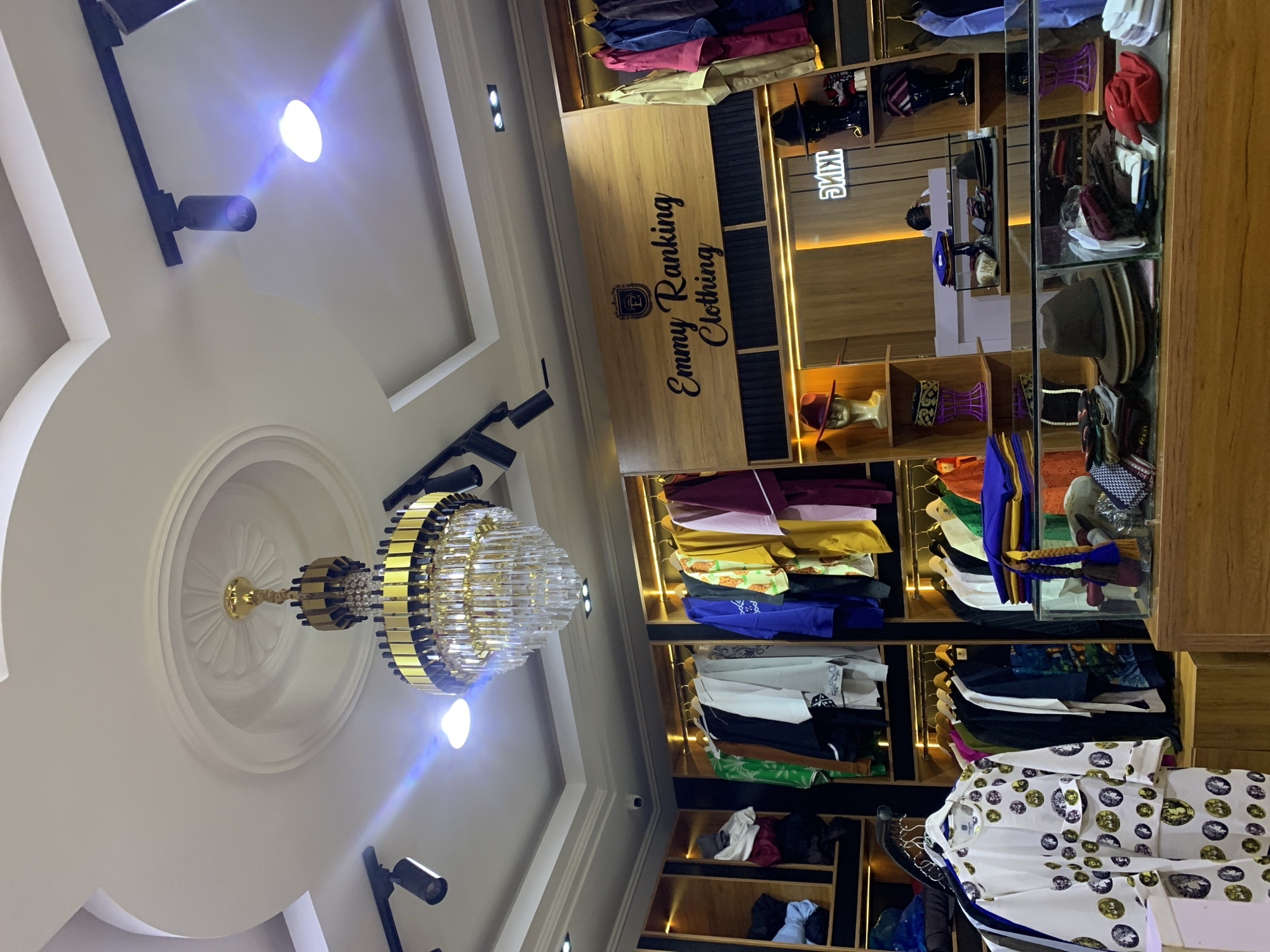 The image shows the interior of a clothing store in Gura Top, Nigeria. The store features a chandelier, track lighting, and wooden shelves displaying various clothing items, including shirts, pants, and accessories. There are mannequins dressed in hats and other headwear. A sign on the wall reads "Emmy Ranking Clothing". A woman is visible in the background, appearing to be an employee or customer. The store appears to be well-organized and stocked with a variety of apparel.