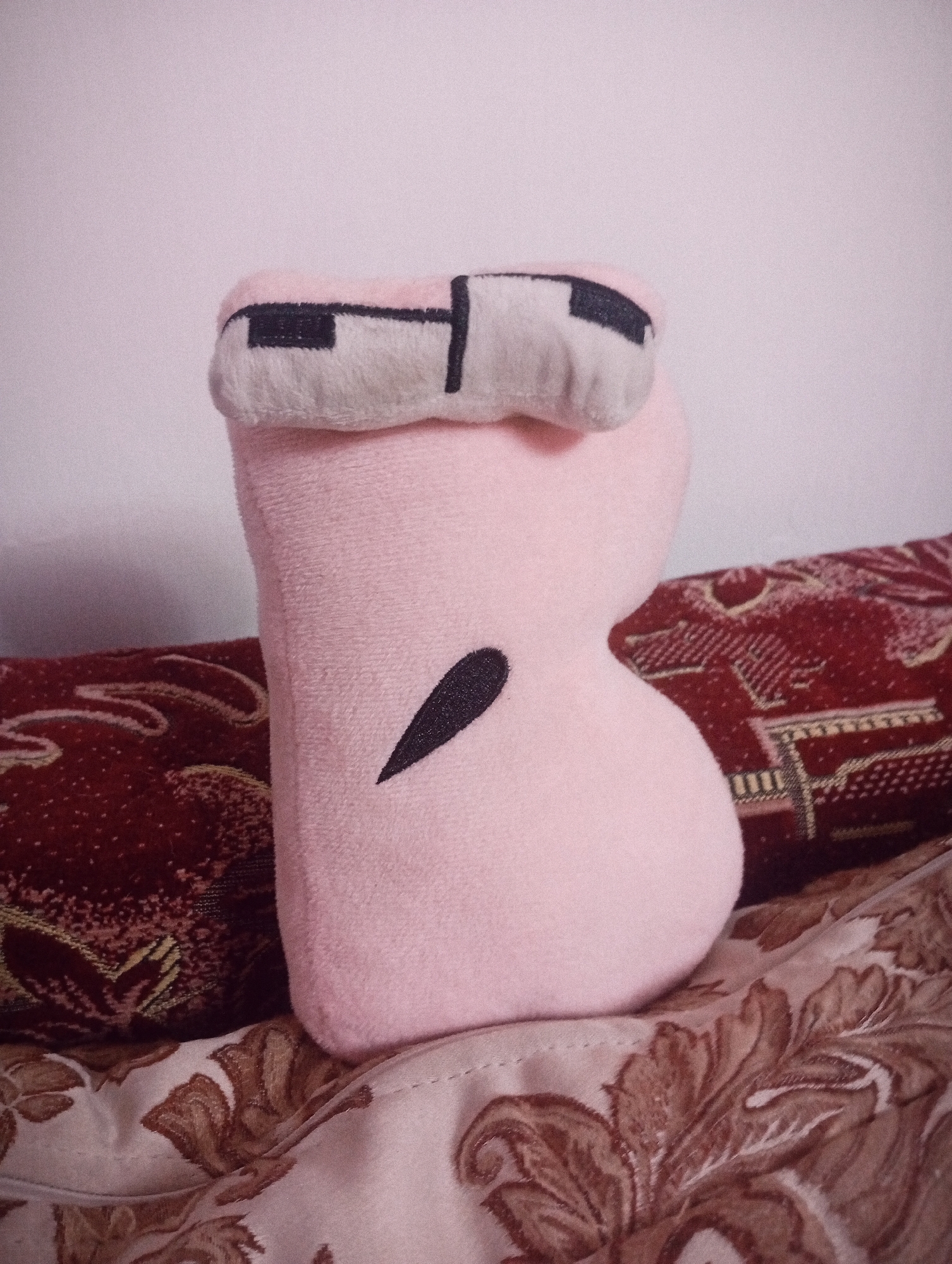 A soft pink plush toy shaped like the letter 'B' is positioned on a surface covered with patterned fabric. The toy has a fuzzy texture and features black stitching to represent facial features: a horizontal line across the top and a curved, pointed shape on the lower curve of the 'B'. Behind the toy, there is a patterned textile with red and gold accents. The background is a plain, light-colored wall. The lighting is soft, and the image appears to be taken indoors. There are no people visible in the image.