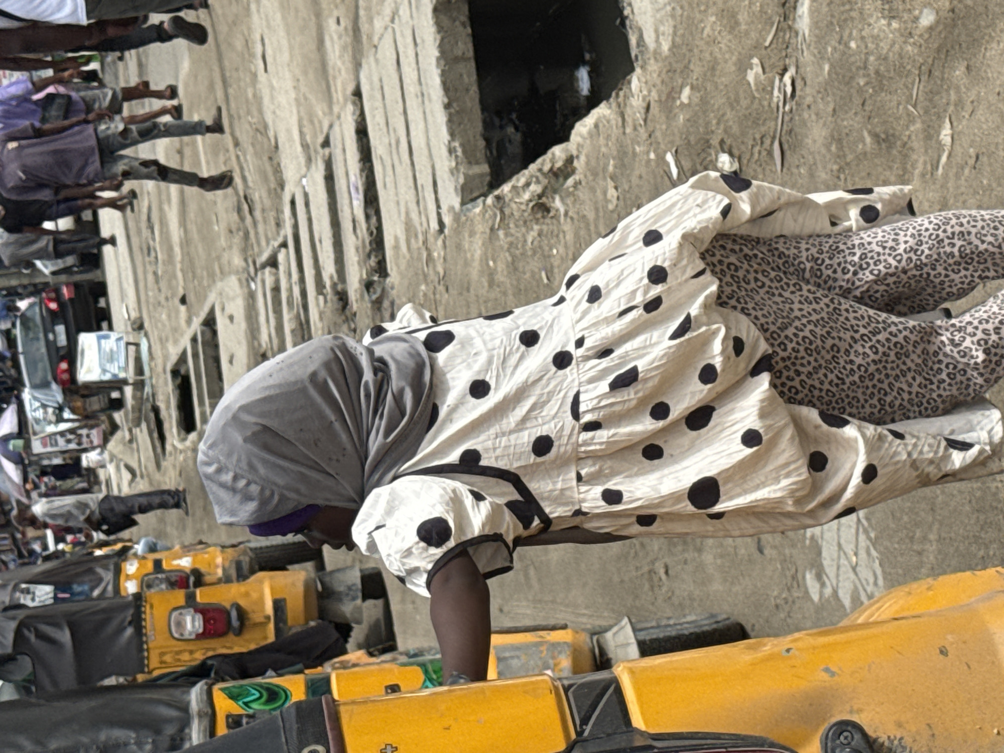 In Lagos, Nigeria, a person wearing a gray head covering, a white polka-dotted dress, and leopard-print leggings is positioned near a row of yellow motorcycles. Several other individuals are visible walking in the background. The setting appears to be an urban street environment.