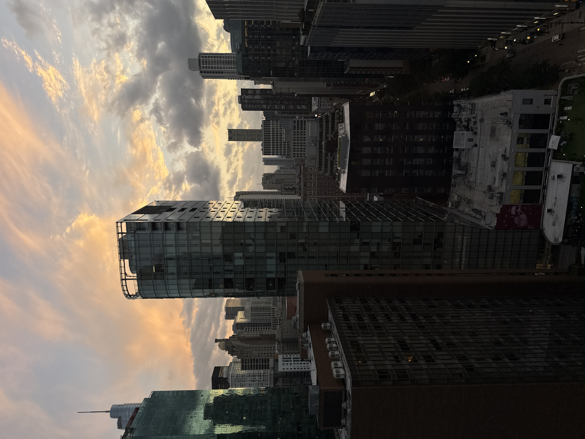 The image captures a dusk view of the Chicago skyline, with a focus on several high-rise buildings. The sky is filled with dramatic clouds tinged with the warm hues of sunset, ranging from soft oranges and yellows to darker grays.
Prominent in the foreground is a dark, brick-faced building with numerous windows and several air conditioning units visible on its upper floors. To its right, a tall, modern skyscraper with a reflective glass facade dominates the view, its structure curving slightly. Further into the cityscape, a dense collection of various buildings, both older and contemporary, rises against the sky. These include structures with intricate architectural details and others with stark, glass exteriors.
The perspective suggests the photo was taken from a high vantage point, likely another building, looking eastward or southeastward over the city. The lighting is dim, characteristic of twilight, with some building windows illuminated, indicating occupancy. The street level is largely obscured by the buildings, but glimpses of streetlights and a few cars are visible, hinting at the urban environment below. No people are clearly discernible in the scene.