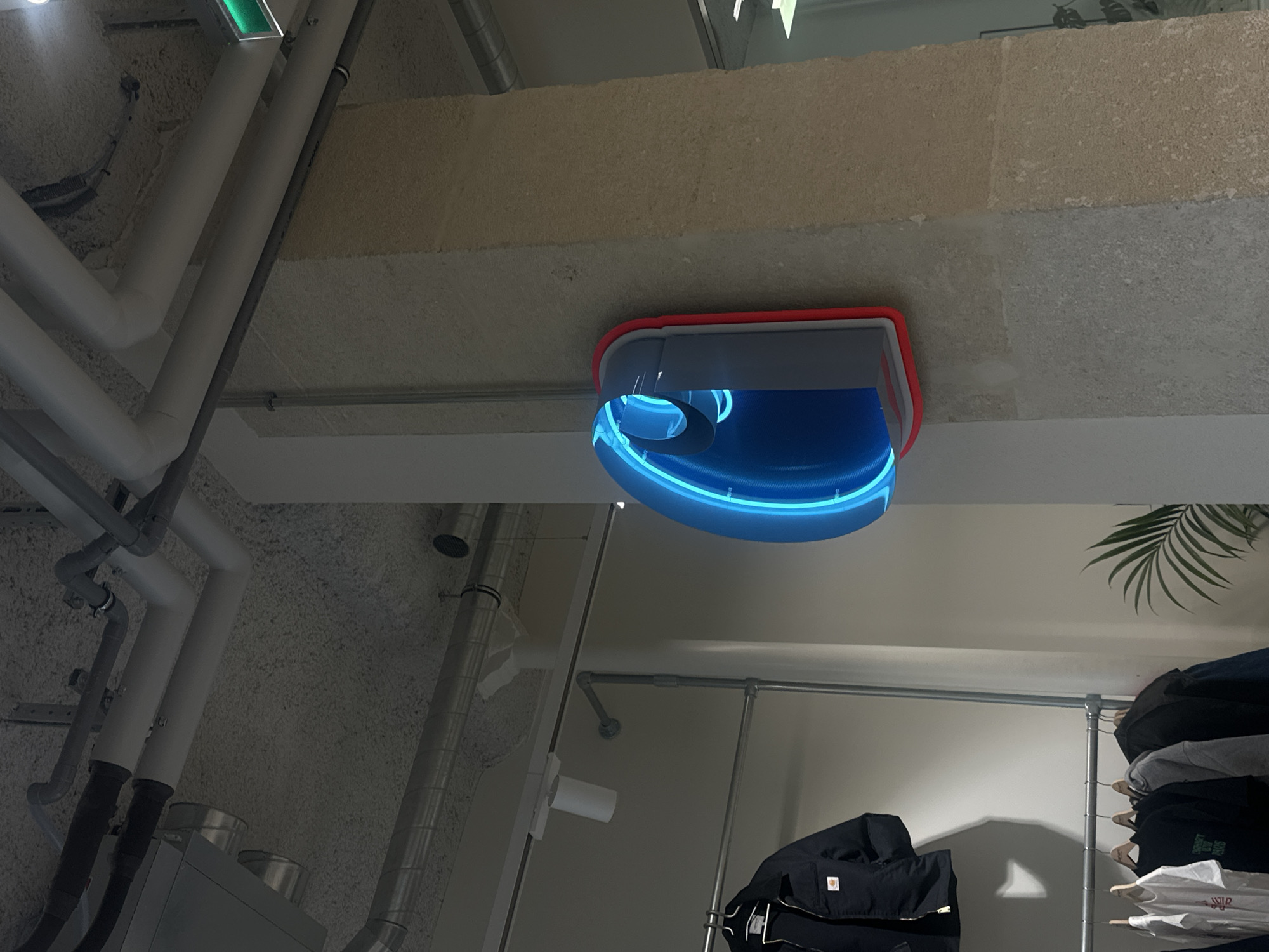 The image depicts an interior space with exposed industrial elements and retail displays.
In the foreground, a complex network of white and gray pipes runs along the ceiling and wall, indicative of an industrial or commercial setting. A lighting fixture with a red and gray frame and blue neon accents is attached to the ceiling.
Below the ceiling, a clothing rack with a variety of garments is visible. The clothes are stacked and hung on hangers, displaying different colors and styles. A green potted plant sits near the rack.
The background is a textured concrete wall. The overall impression is a modern, urban interior with a blend of industrial design and retail display. There is no verifiable evidence of a location within Paris, France.