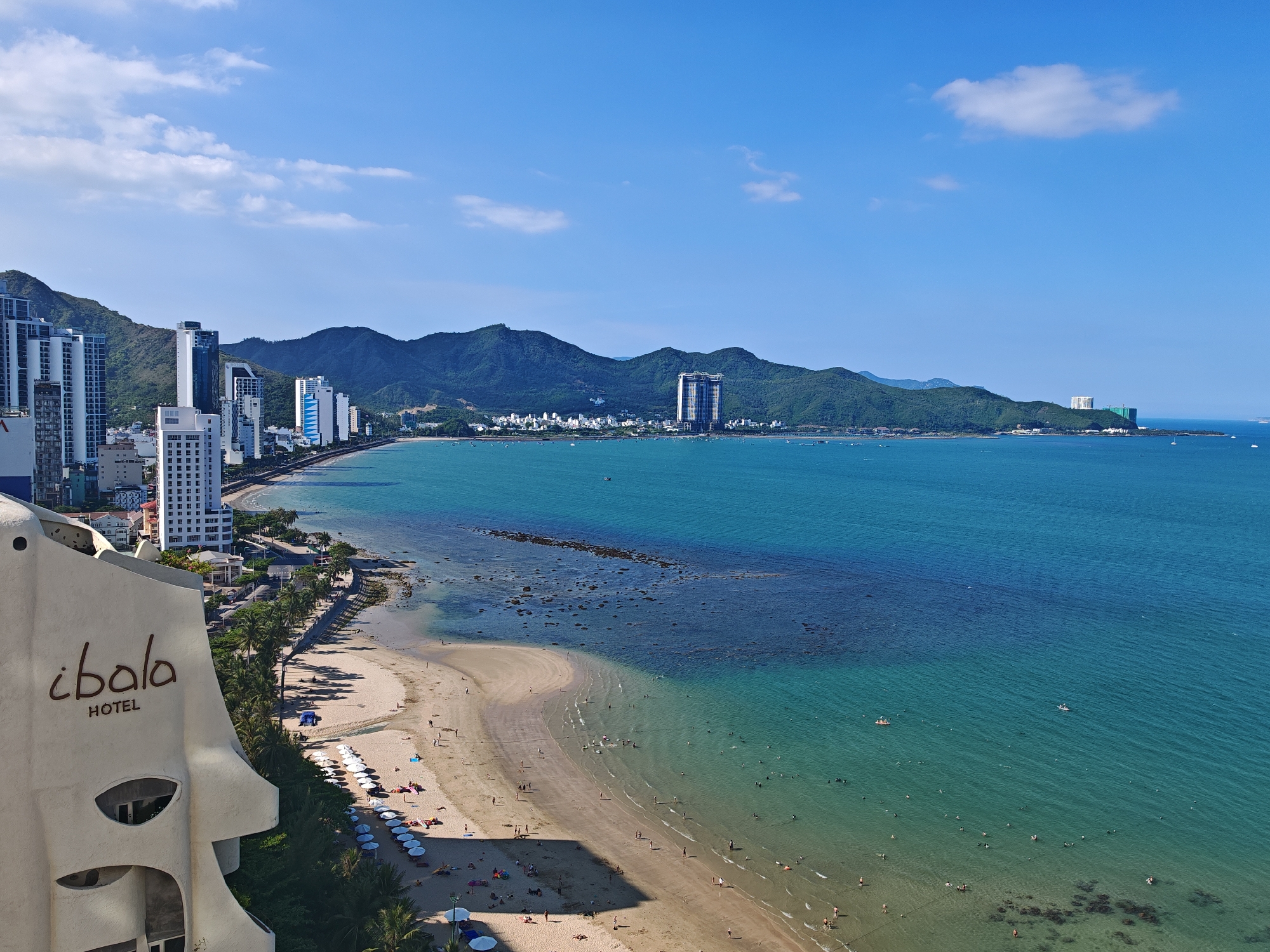 This is a high-angle, wide shot of a coastal city, likely Nha Trang, Vietnam, on a bright, sunny day with a clear blue sky and scattered white clouds. The foreground features the distinctive, pale beige, curved facade of the "i Bala HOTEL" with its logo prominently displayed. Adjacent to the hotel is a sandy beach lined with palm trees and numerous white umbrellas, beneath which sunbathers are visible. A calm, turquoise ocean stretches out to the horizon, dotted with small boats and many people swimming and wading near the shore. In the mid-ground, a coastline curves into the distance, bordered by a road and a row of tall, modern buildings, including hotels and apartments, interspersed with lush green hills and mountains. The water exhibits varying shades of blue and turquoise, with visible rocky formations and sandbars in shallower areas. The overall scene depicts a bustling beach resort environment.