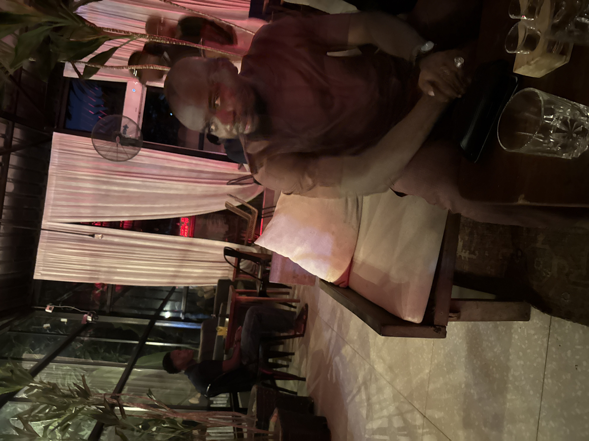 The image depicts an indoor section of a lounge or restaurant, located in Abuja, Nigeria, during nighttime hours, characterized by dim ambient lighting.
In the immediate foreground, positioned to the right, a dark-skinned man with a bald head and a beard is seated on a light-cushioned, dark wooden bench. He wears a solid dark reddish-brown short-sleeved shirt and matching trousers. His hands are clasped together on a dark wooden table directly in front of him; a watch is visible on his left wrist and a ring on his right hand. On the table, there is a dark rectangular object, a clear patterned drinking glass, and two smaller clear glasses resting on a small wooden block.
In the mid-ground, to the left, a second dark-skinned man is seated on a black chair at a small, dark wooden table. He is dressed in a dark short-sleeved shirt and dark trousers, wearing sandals on his feet. He faces towards the right.
The background features tall glass paneling with dark frames, through which additional blurred figures are discernible. Floor-length white curtains are draped along a section of the back wall. Above the curtains on the right, a black wall-mounted fan is visible, positioned above an air conditioning unit. Large potted plants with green foliage are present, one prominently on the far left and another partially on the right. The ceiling is dark, and the light-colored floor appears tiled, exhibiting shadows. Distant red lighting is faintly visible through the glass and curtains in the far background. No direct interactions between the visible individuals are evident.