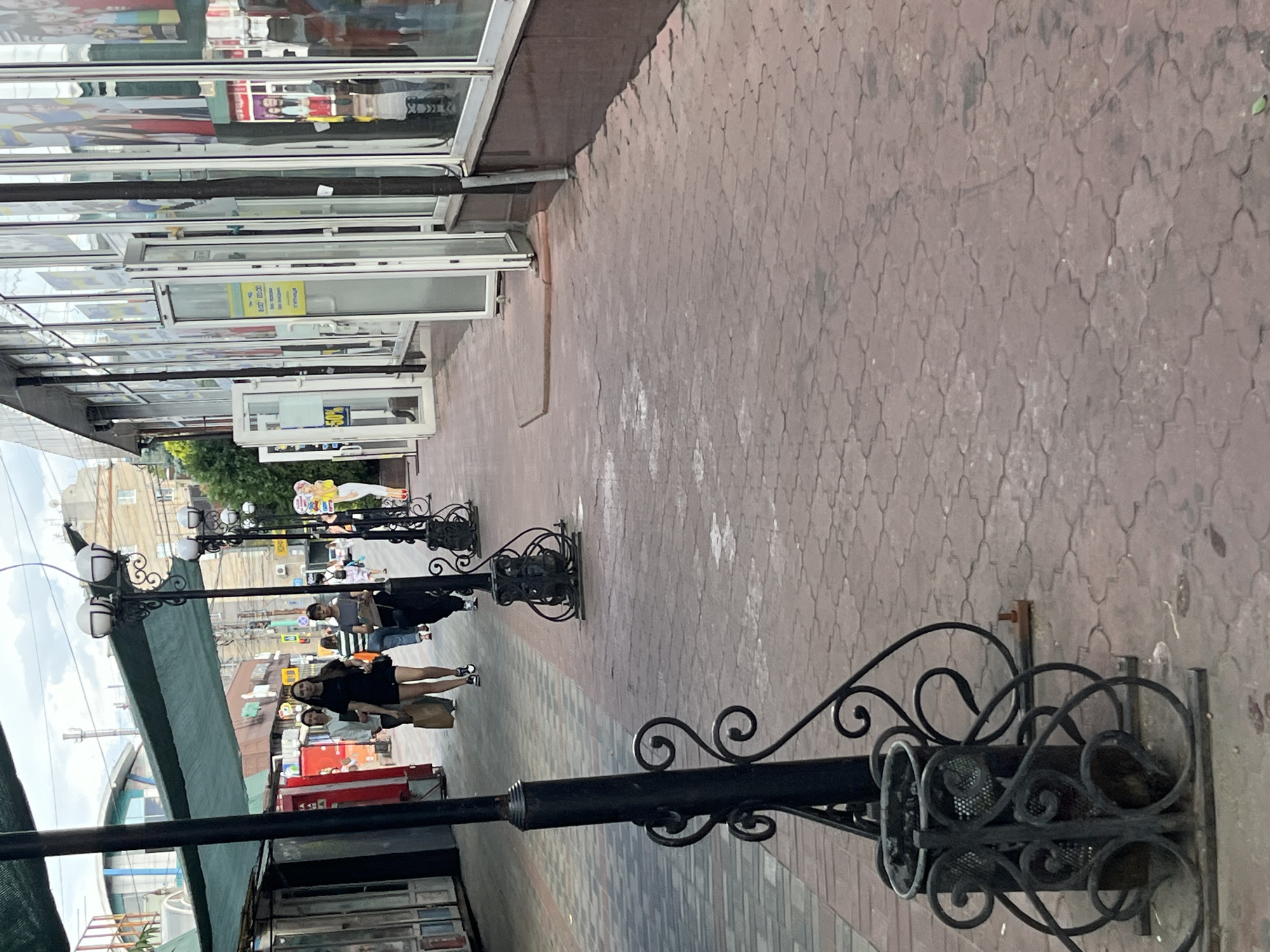 This media file depicts a street scene in Dnipro, Ukraine, likely during the daytime given the lighting. The perspective is from the sidewalk, looking down a street lined with shops on the left. The ground is paved with reddish-brown interlocking bricks arranged in a decorative pattern.
Several people are visible walking along the sidewalk. On the far left, there are storefronts with large glass windows, reflecting the surroundings and displaying various items, though the details within are somewhat obscured. Further down the street, more individuals are walking, some appearing to be engaged in conversation or shopping.
Lining the sidewalk are decorative black lampposts, some with multiple lights. The lampposts have ornate metalwork, including scroll designs and what appear to be basket-like structures at the base of some.
In the background, buildings can be seen, suggesting an urban or commercial area. The weather appears to be overcast or hazy, as the light is diffused and there are no harsh shadows.
There is some visible text on signs within the shop windows, such as a yellow sign with Cyrillic text that might be an advertisement or informational notice. Another sign in a window on the right appears to have "50%" on it, possibly indicating a sale. The overall atmosphere suggests a typical day in a pedestrian-friendly urban environment.