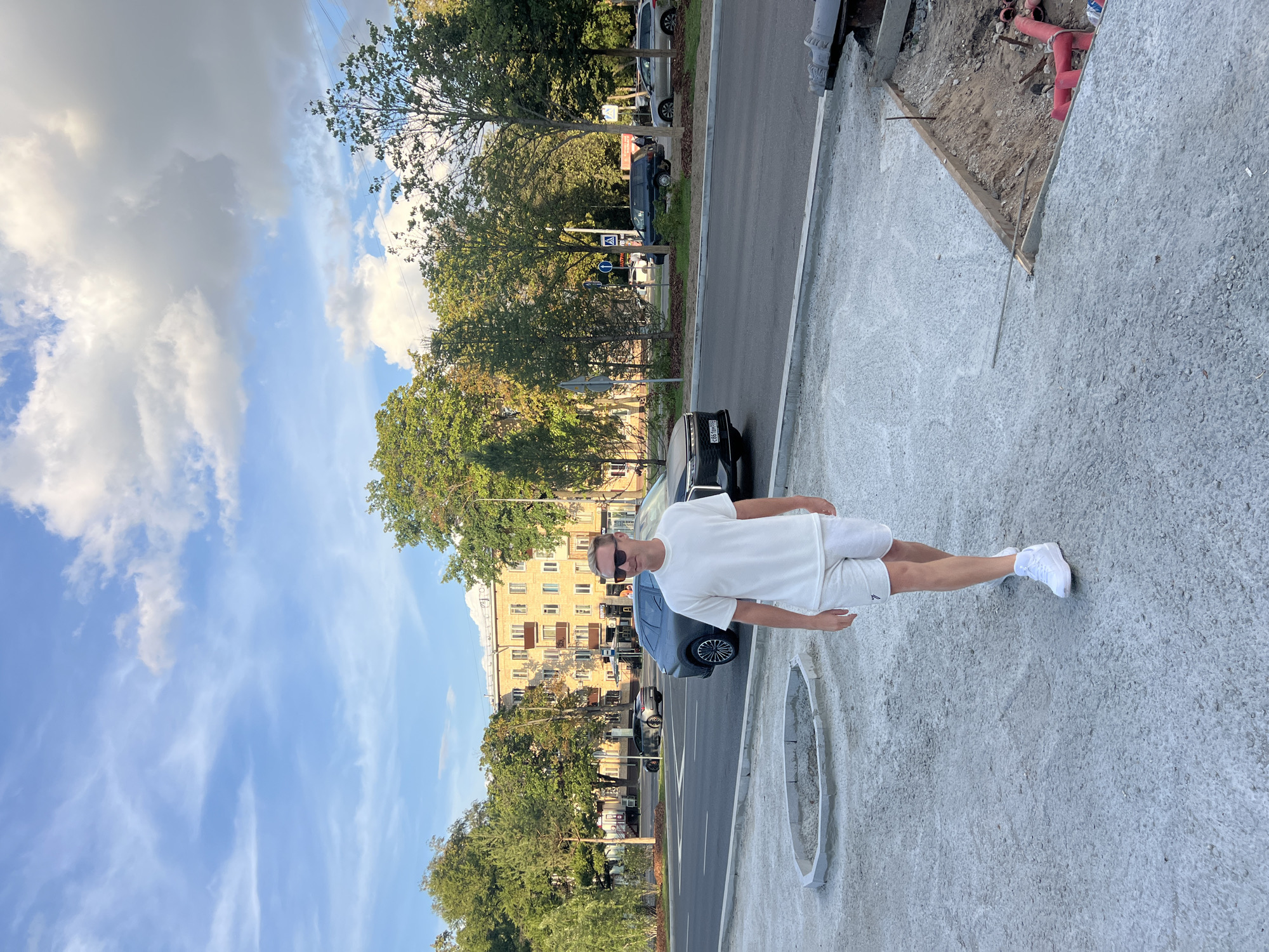 A light-skinned man wearing sunglasses, a white t-shirt, and white shorts walks towards the camera on a construction site sidewalk. Behind him, a street with several cars and a multi-story building is visible. Trees line the street, and the sky is partly cloudy. The image appears to be taken in Moscow, Russia, based on the Cyrillic script visible on a street sign.