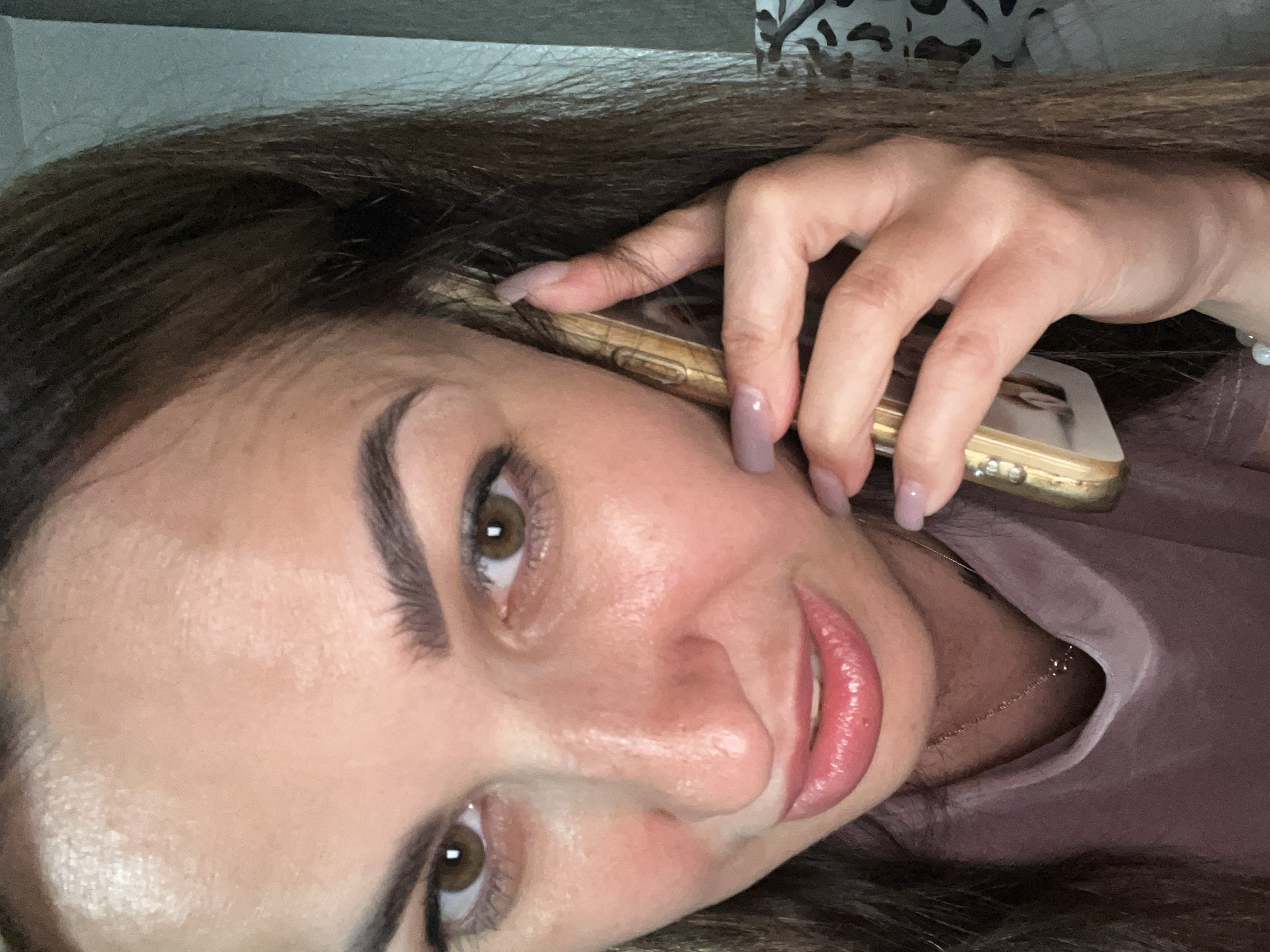 A medium shot shows a woman holding a smartphone to her ear with her right hand, looking at the camera with a slight grimace. Her eyes are brown, and she is wearing makeup. Her lips are full and painted with a glossy pink lipstick. She has long, dark brown hair that falls around her shoulders. The background is blurry, but appears to be an indoor setting with a patterned textile visible on the right. The lighting is soft and appears to be coming from the front. The woman is wearing a pale pink garment that is partially visible. The smartphone case is gold and has some decorative elements.