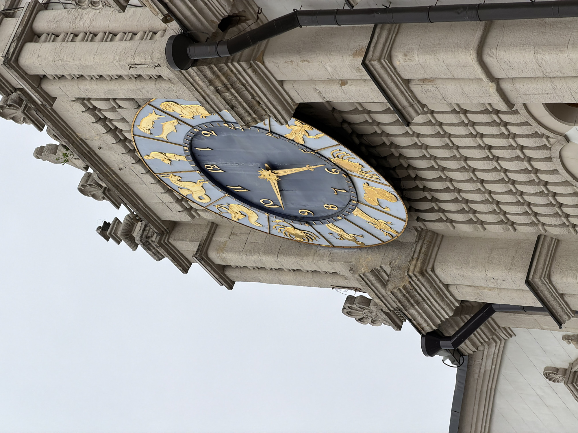 The image depicts a clock mounted on the exterior of a building. The clock face is oval-shaped with a dark blue background and gold numerals and zodiac symbols around the perimeter. Gold hands indicate the time. The building is constructed of light-colored stone with ornate carvings and moldings. A black drainpipe runs along the top of the building, near the clock. The background is a gray, overcast sky. This is the clock face of the State History Museum, Moscow, Russia.