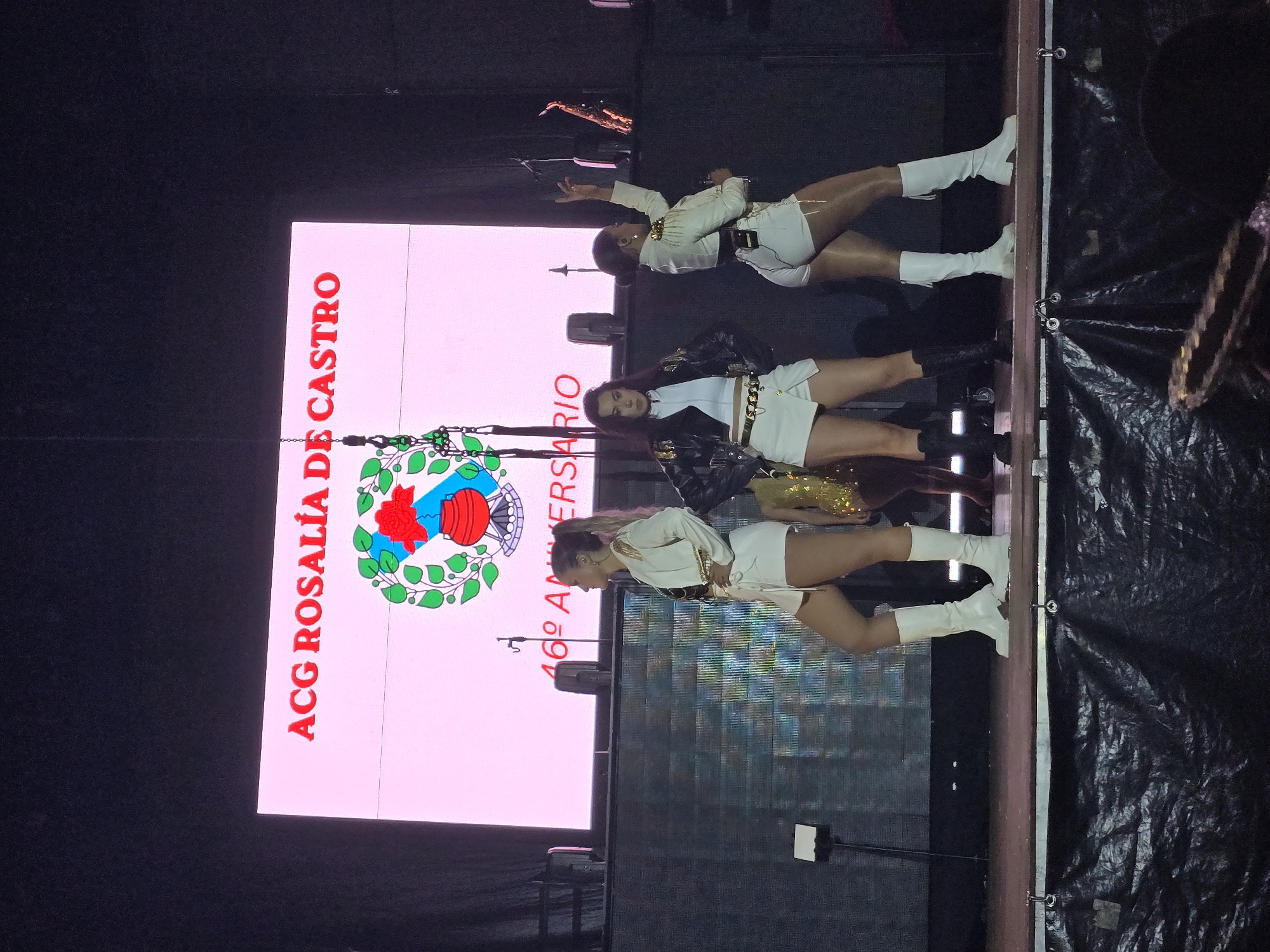 A stage performance is captured in an unspecified venue within Cornellà de Llobregat, Spain. The background is dominated by a large, brightly lit LED screen displaying "ACG ROSALÍA DE CASTRO" in red capital letters at the top. Below this text, a circular emblem features a green wreath, a blue shape resembling a map or coat of arms, a red stylized flower or crest, and a red pot. Beneath the emblem, "46º ANIVERSARIO" is displayed in red text. The screen emits a pinkish-white light.
Three female performers are positioned on a dark brown stage. The stage front is covered with a black, textured material, possibly a tarp or fabric.
1. **Left performer:** Stands in profile, facing right, with light-colored hair in a high ponytail. She wears a white, long-sleeved blazer-style dress or romper with gold-colored shoulder embellishments and a matching belt. White knee-high boots complete her attire. Her left hand rests on her hip.
2. **Center performer:** Stands facing forward, slightly angled towards the viewer's left, with dark hair. She wears a black leather jacket over a white top, white shorts, and black ankle boots. A gold-colored chain belt is visible at her waist. Her hands are placed on her hips.
3. **Right performer:** Stands with her back partially to the viewer, turned slightly to her right, with dark hair in a bun. She wears a white, long-sleeved blazer-style dress or romper, similar to the left performer, with gold-colored shoulder embellishments and a matching belt. White knee-high boots are worn. Her right arm is raised above her head.
Additional stage elements include a black microphone stand with a white tablet holder to the left of the left performer, several dark speakers or light fixtures behind the performers, and a gold-colored saxophone on a stand on the far right of the stage. A heavy-duty chain with a hook hangs vertically from above, partially obscuring the screen's lower left area. The stage is brightly illuminated, casting shadows beneath the performers. A blurred, dark object is visible in the bottom right corner of the frame.