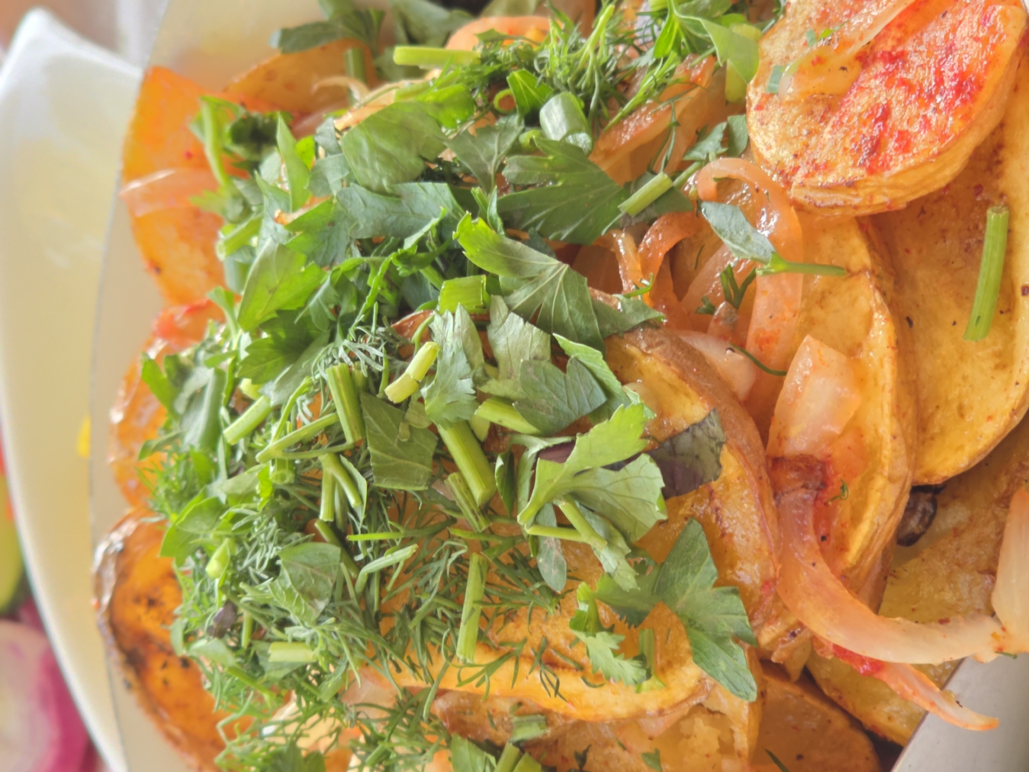 The image shows a close-up of a dish of roasted potatoes and onions, topped with fresh green herbs. The potatoes are cut into wedges and appear to be seasoned with paprika, giving them a reddish-orange hue. The onions are thinly sliced and translucent. The herbs, including dill and parsley, are finely chopped and generously scattered over the dish. The overall presentation suggests a rustic and flavorful meal. The image is taken from a slightly elevated angle, focusing on the texture and colors of the food. There is no indication of the city of Leninakan, Russia, in the image.