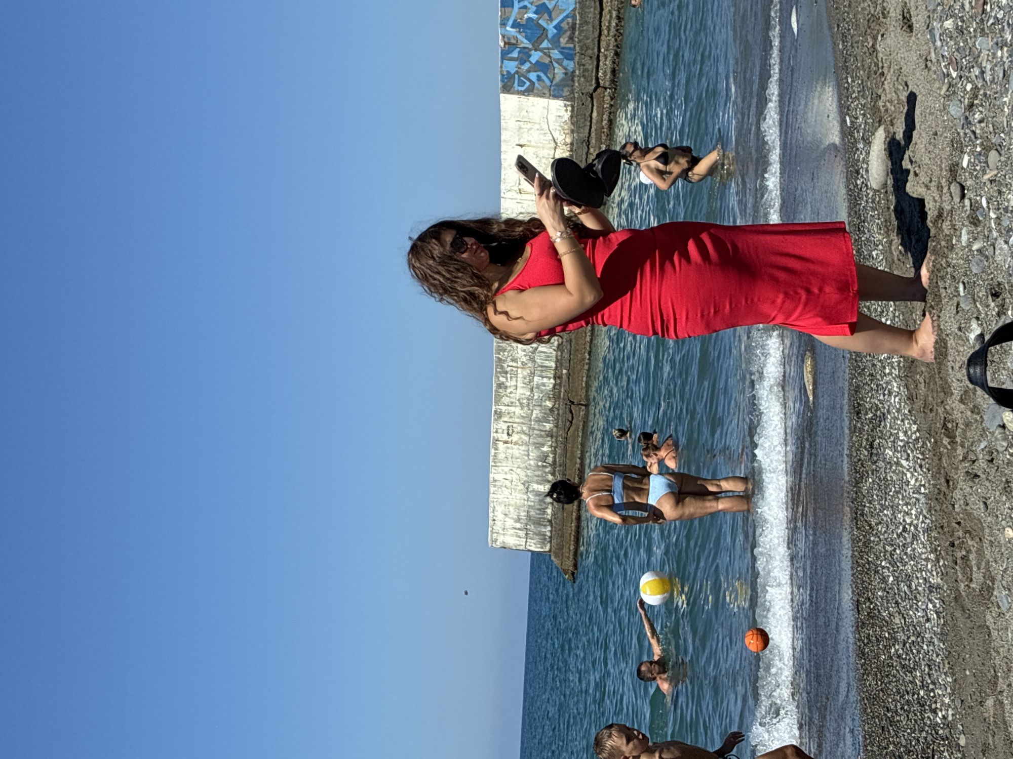 In Sochi, Russia, on a sunny day, people are enjoying the Black Sea. A woman in a vibrant red dress stands on the pebbled shore, looking at her phone and holding a pair of black flip-flops. In the water, several individuals are swimming and playing. A man in the background is reaching for a yellow and white beach ball, while a young boy is partially visible on the left side of the frame. Further out, other swimmers are dispersed in the blue water. A concrete pier, partially covered with graffiti, juts out into the sea in the background. The sky is a clear, bright blue.