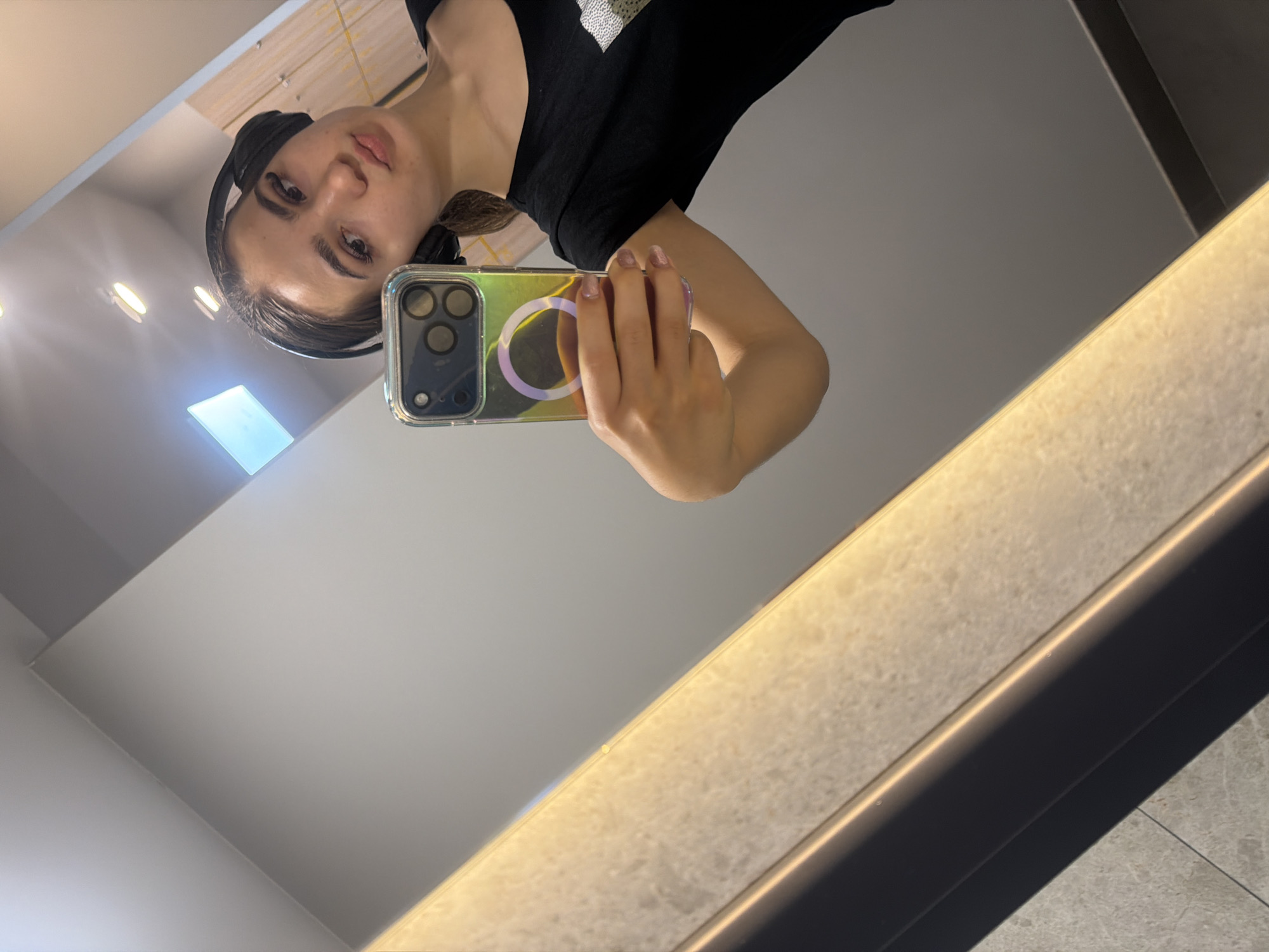 A selfie is reflected in a mirror. A young woman with headphones poses holding a cellphone with a clear case. The surrounding environment appears to be an interior space with neutral-colored walls. The city of Sofia, Bulgaria cannot be determined from the image.