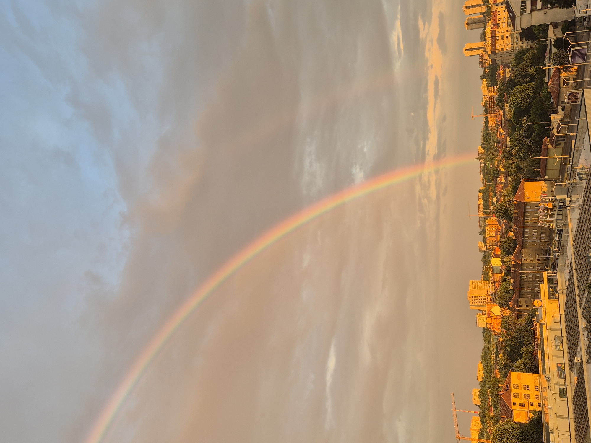 This image captures an urban landscape in the city of Unknown, Unknown, bathed in the warm, golden light characteristic of either sunrise or sunset. The sky, occupying the upper two-thirds of the frame, features a mix of pale blue in the top right corner and extensive, diffused clouds predominantly colored with warm orange, pink, and grey hues. A prominent, vibrant primary rainbow arches diagonally across the sky from the lower left, extending upwards and rightwards, displaying a full spectrum of colors. A fainter, secondary rainbow is discernable above and outside the primary one, particularly in the upper middle section of the sky.
The scene below the horizon line depicts a dense collection of buildings, trees, and urban infrastructure. In the midground, various multi-story residential and commercial buildings with both pitched and flat roofs are visible. One notable structure in the lower center features a flat roof with a visible "LIDL" sign and an adjacent parking lot containing several parked cars. Abundant green foliage from numerous trees is interspersed among the buildings, especially on a sloping hill in the background. Several construction cranes, including a prominent yellow one on the far left and others scattered towards the center-right, indicate ongoing development within the city. Taller, more modern apartment blocks are visible on the distant horizon, particularly towards the right side. The overall illumination casts long shadows and highlights building facades with a strong golden glow. No individuals are explicitly discernible within the frame.