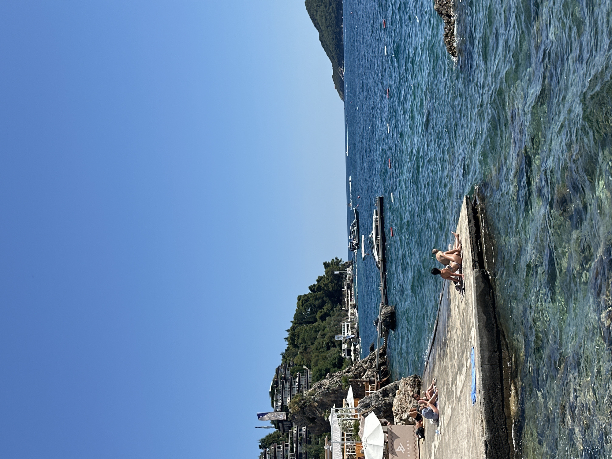 This image depicts a coastal scene in Budva, Montenegro, likely from the Mogren Beach area, on a clear, sunny day. The foreground features a concrete pier with several individuals sunbathing. Two people, a man and a woman, are seated facing the sea on the right side of the pier. Further to the left, another group of people are reclining on towels. The water is a vibrant blue, with small waves rippling the surface. Beyond the pier, the sea extends to the horizon, with a few boats visible, including speedboats and yachts. The coastline is characterized by a rocky incline densely covered in lush green trees. Perched on the hillside are buildings with multiple levels, appearing to be hotels or resorts, with balconies and terraces overlooking the sea. White umbrellas and structures, possibly part of a beach club or restaurant, are visible along the shore. In the distance, a verdant island or peninsula rises from the sea. The sky is a clear, bright blue, with no clouds present.