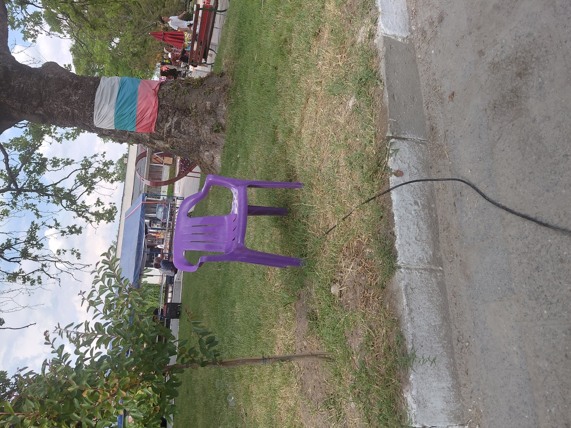 The image depicts an outdoor public area in Kostievo, Bulgaria, likely a park or market setting, under a partly cloudy sky.
In the immediate foreground, a purple plastic chair with armrests stands on a patch of grass. To its left, a slender young tree with green foliage is visible. A dark, possibly electrical, cable runs across the lower asphalt ground from the left edge towards the lower right. A concrete curb separates the asphalt from the grass.
The midground features a large, mature tree with a thick, textured trunk, around which a fabric strip displaying the colors white, green, and red (consistent with the Bulgarian flag) is wrapped horizontally. To the right of this tree, multiple people are present, including individuals walking and standing near what appear to be market stalls or vendor setups with red umbrellas and white tables. One person in a white shirt and dark pants is walking to the right, while others are less distinctly visible. A wooden bench is situated near these stalls. To the left of the large tree, a blue tent and another white structure are visible, with further structures and possibly vehicles in the background. A decorative heart-shaped planter is also near the base of the large tree.
The background shows additional green trees and shrubs, along with a light-colored building with a blue roof visible beneath the sky. The overall ground covering alternates between asphalt, concrete, and varied grass patches, some appearing dry or sparse.