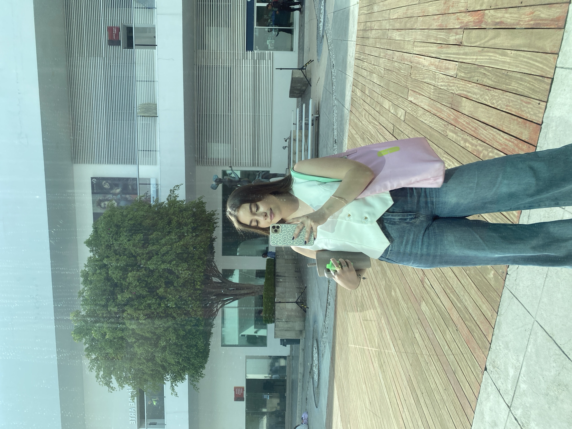 A woman is taking a selfie in front of a glass window reflecting a building and a tree. The building has multiple windows and horizontal architectural elements. The woman is wearing a white top, blue jeans, and a pink bag. She is holding a phone in one hand and a cylindrical object in the other. The ground is made of wooden planks and concrete tiles. The city is unknown.