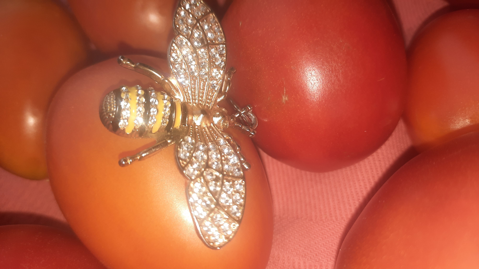 A close-up shot shows a gold-colored bee brooch adorned with small clear stones, placed on top of a cluster of tomatoes. The brooch is positioned on one of the larger, orange-hued tomatoes, with its wings spread and legs slightly extended. The tomatoes are a mix of orange and red, with some of the red ones appearing closer to the foreground. The background is a soft pink fabric. There is no indication of the location being in the city of Шклоў, Belarus.