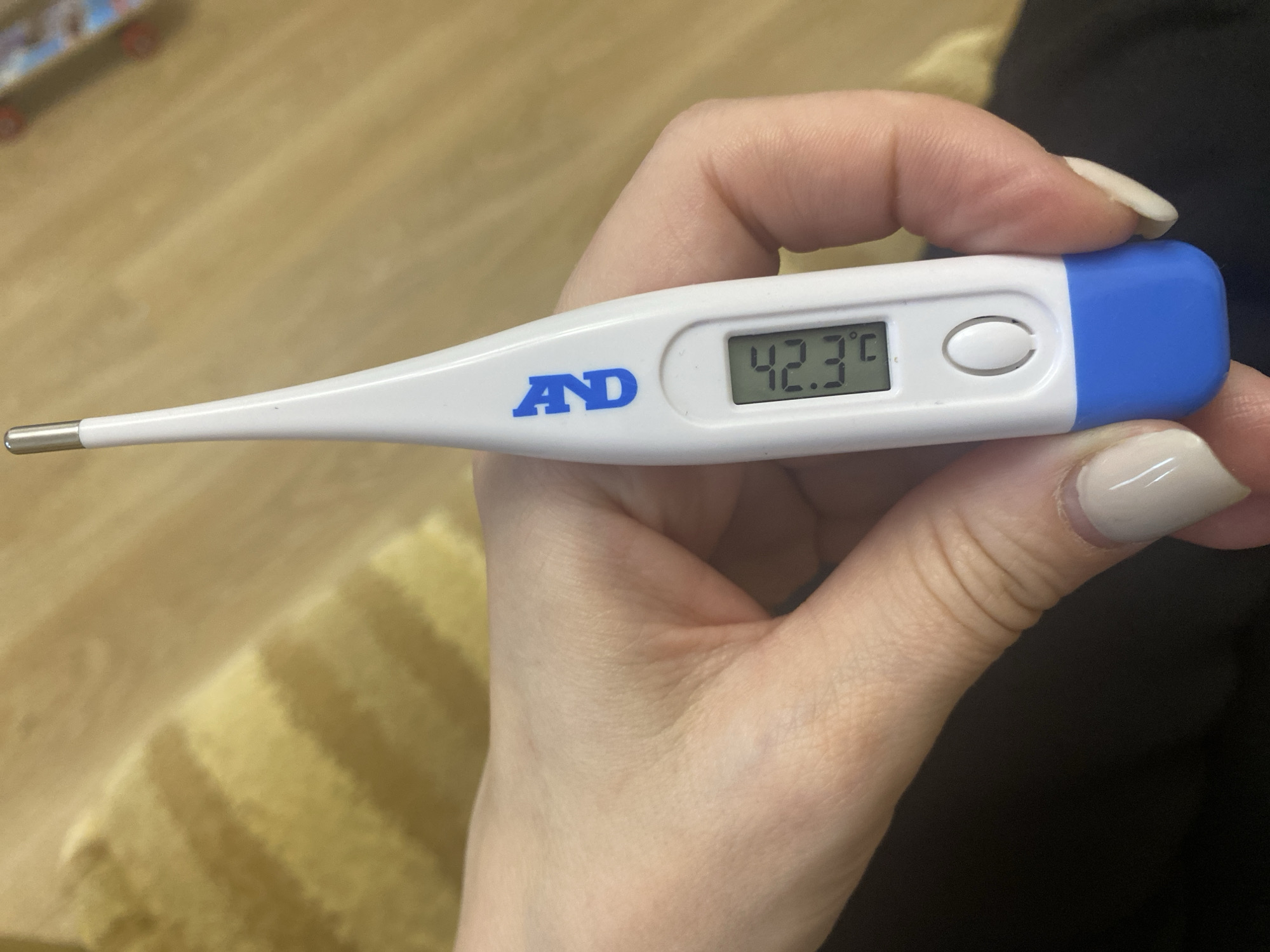 A close-up, horizontal shot features a person's right hand holding a white and blue digital thermometer. The hand has light-colored, manicured nails. The thermometer clearly displays a reading of 42.3°C on its LCD screen. The brand "AND" is visible in blue letters on its white body, which also includes a white oval button near the display. The device has a silver metallic tip at one end and a blue cap at the other.
The background is softly blurred, indicating an indoor setting. It shows a light-colored wooden floor and parts of a striped yellow and brown textile, possibly a rug or blanket. A dark fabric, likely clothing, is also visible in the lower right portion of the image. The activity depicted is the act of taking a temperature, suggesting health monitoring or illness. The reading of 42.3°C is a notably high temperature, indicating a significant fever.