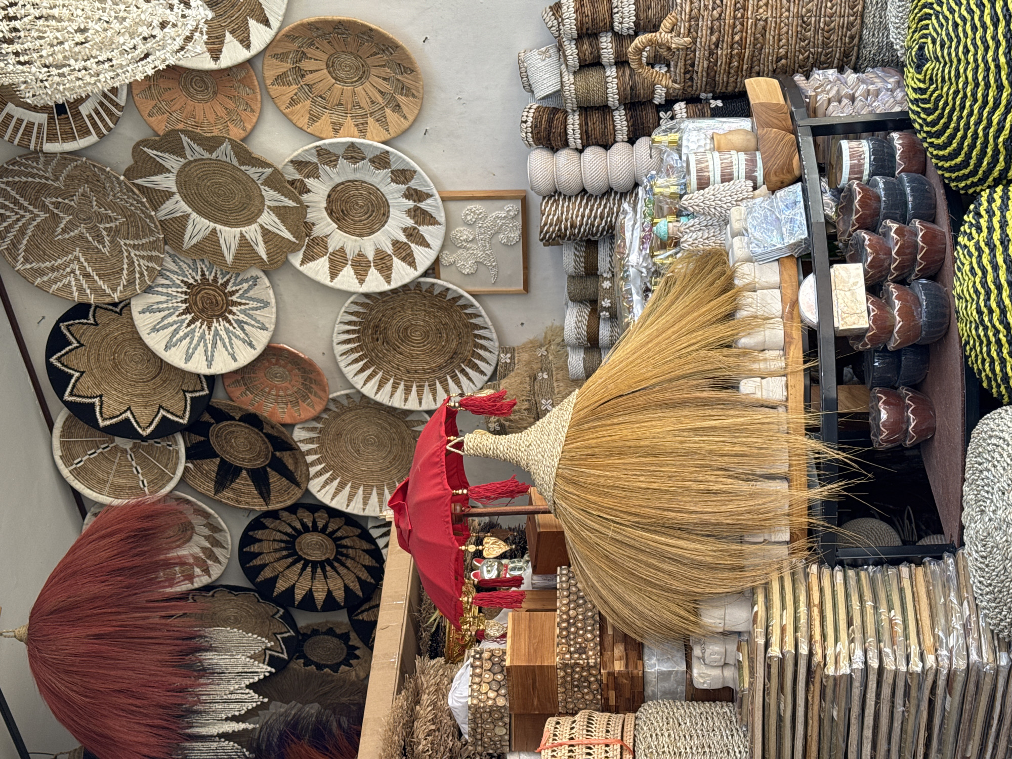 This is a medium shot of a street stall in Ubud District, Indonesia, filled with handcrafted woven goods. The stall is packed with an array of items, predominantly made of natural fibers like straw and rattan. Dominating the center is a large, straw-like decoration with a red umbrella-like top adorned with red tassels and what appears to be a small golden cat figurine. Hanging from the ceiling to the left is a decorative lampshade or ceiling ornament made of red fibers and white beads.
The background is filled with an assortment of round, woven wall hangings, many featuring intricate sunburst or floral patterns in natural, black, and white tones. Below these, the stall displays various wooden boxes, stacked neatly. Further to the right, shelves are laden with smaller woven baskets, some decorated with seashells. In the foreground, stacks of flat, packaged items are visible, and towards the bottom, woven mats with yellow and black stripes are displayed. The scene appears to be a retail environment, possibly a market or souvenir shop, with no people visible. The lighting suggests it is daytime.