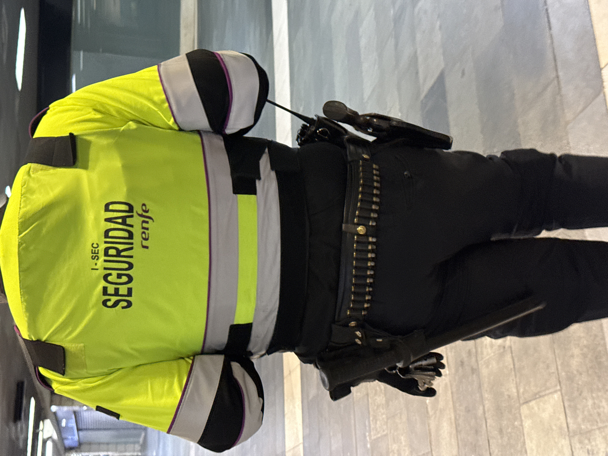 The image shows the back of a security guard wearing a bright yellow jacket with "SEGURIDAD renfe" printed on it. He is also wearing a black belt with ammunition cartridges, a gun holster, and a baton holster. The background appears to be a modern, tiled floor, possibly in a public transport area, such as a train station. The location is likely in Barcelona, Spain, given the "renfe" marking, which is the Spanish national railway company.